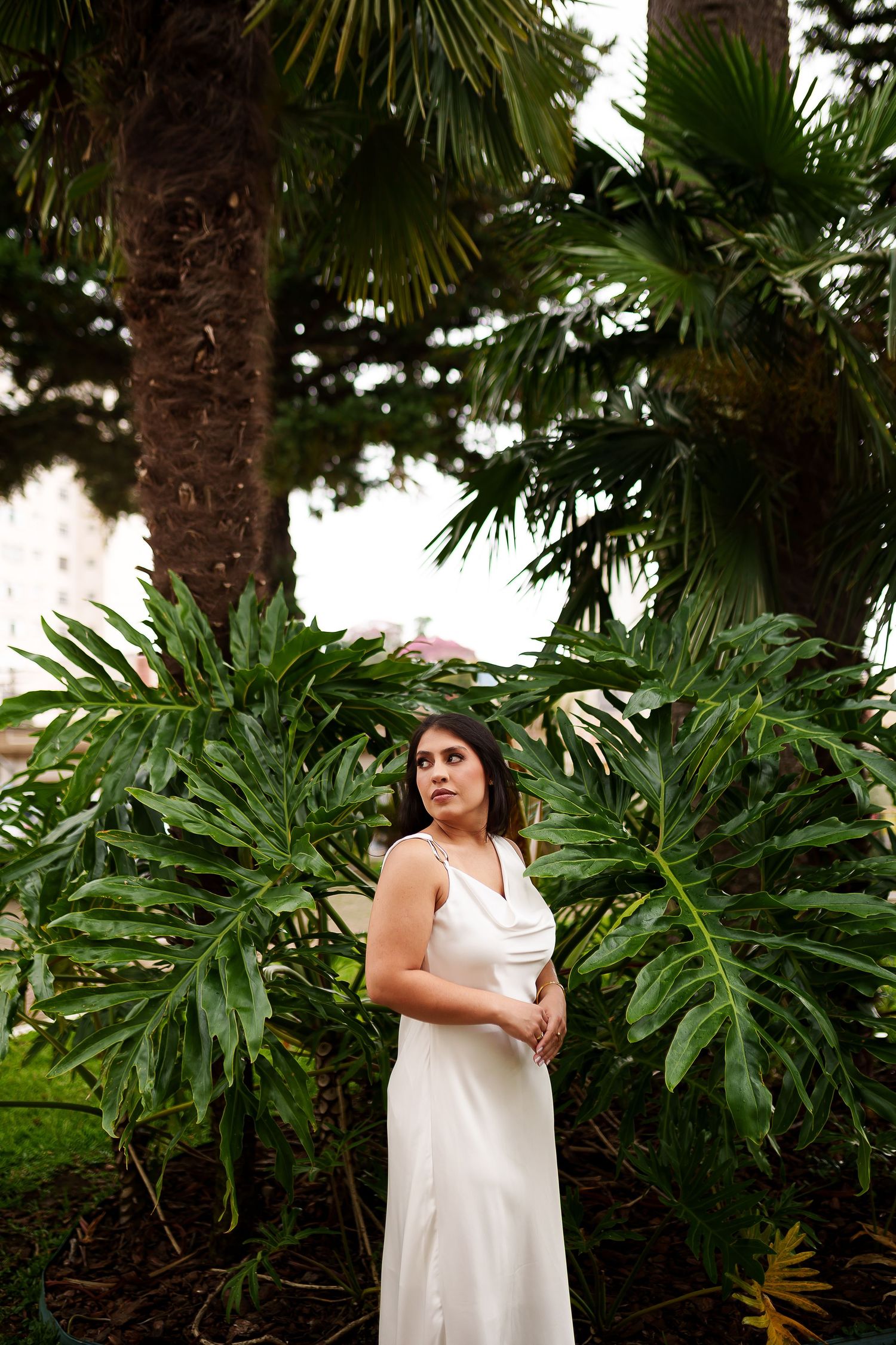 Woman in white dress standing among tropical palm trees and large green monstera leaves in an outdoor garden setting.