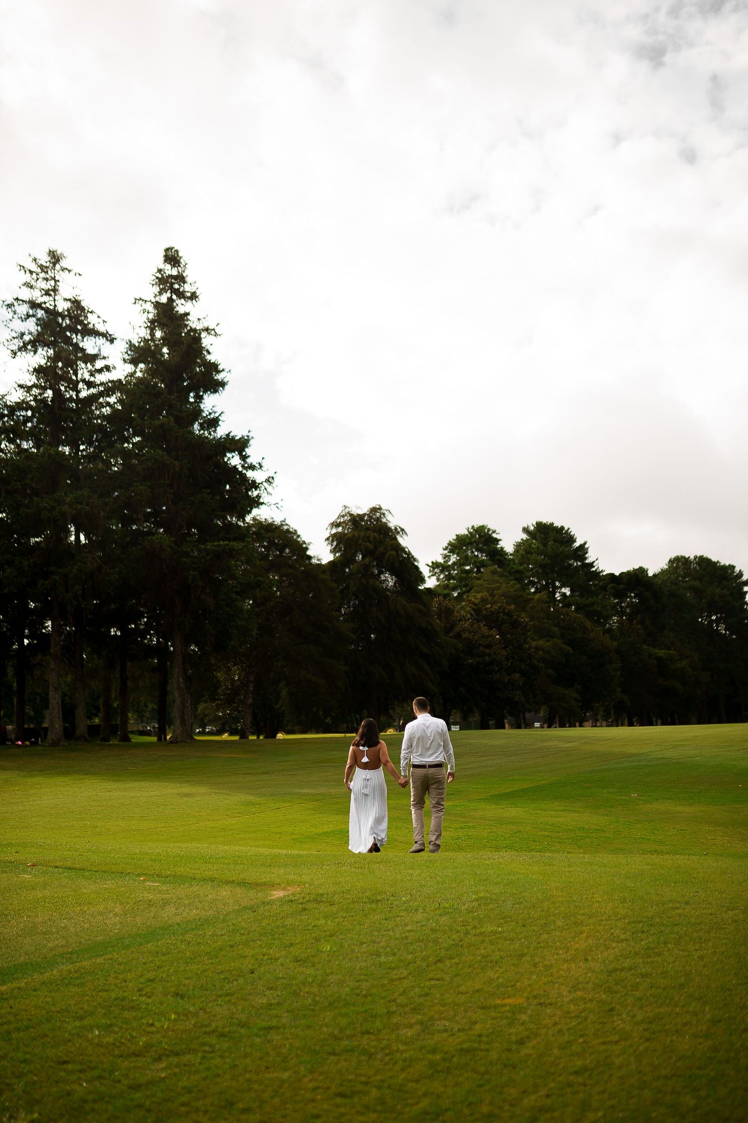 Couple in wedding attire holding hands walking across lush green golf course with tall trees in background.