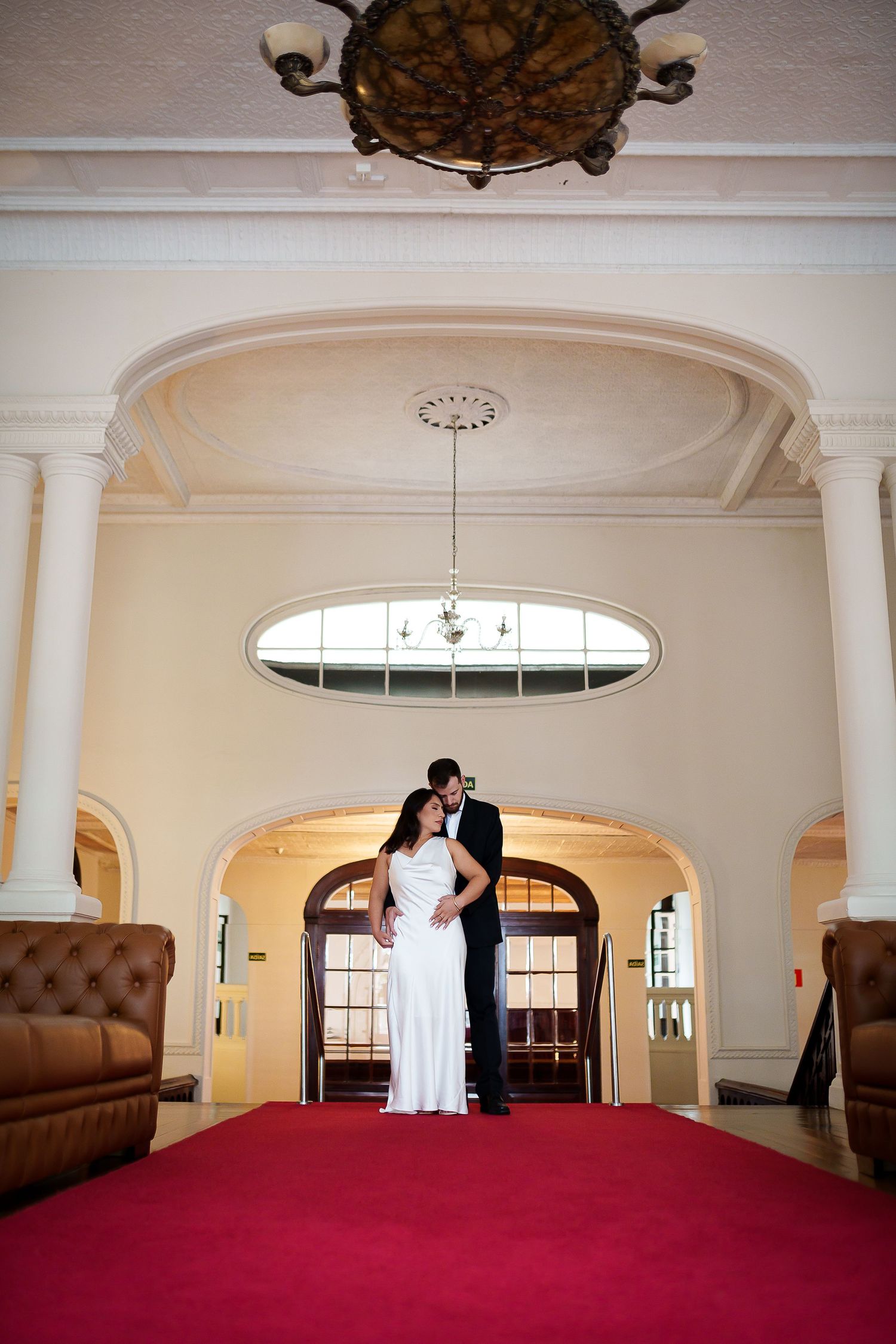 Bride and groom share a romantic kiss on a red carpet in an elegant venue with arched doorways and ornate chandelier.