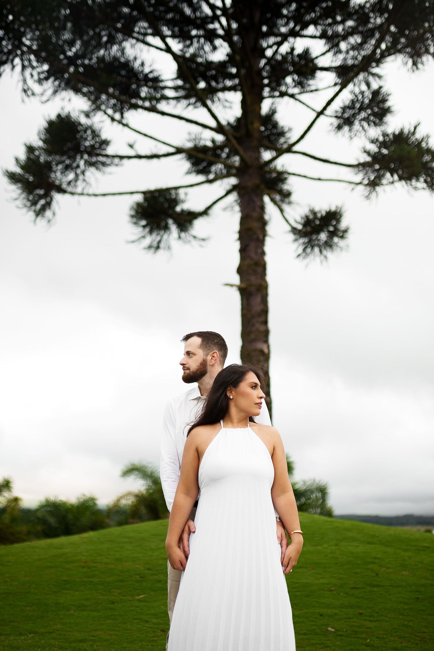 Couple in white attire posing before a tall araucaria tree on a lush green lawn under overcast skies.