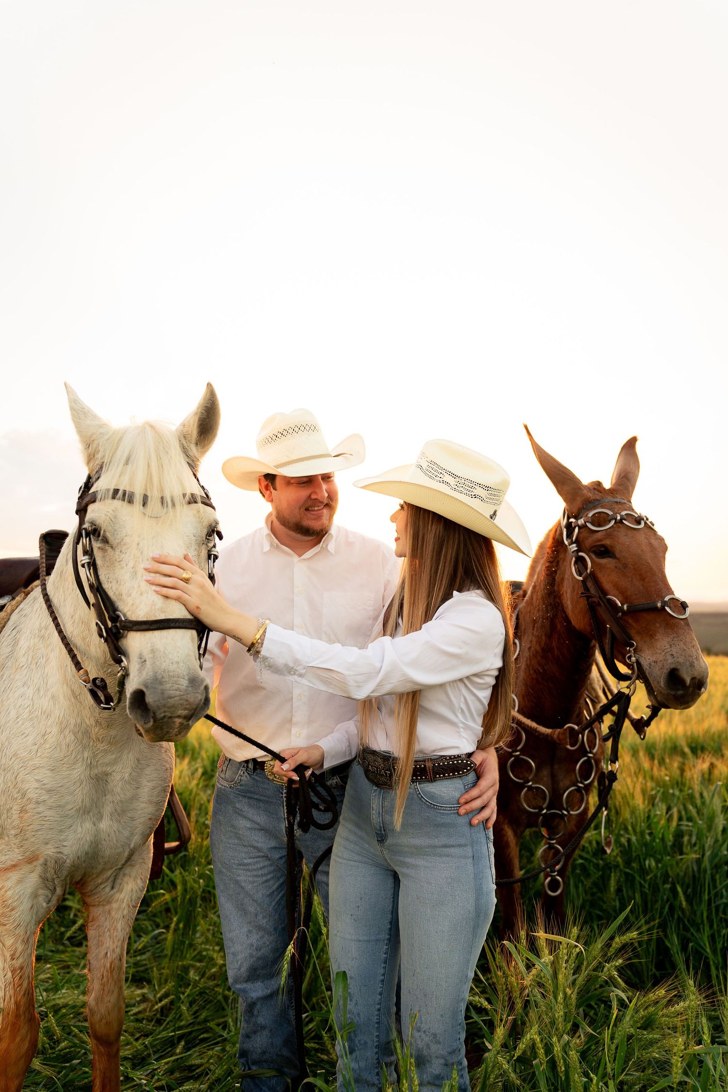Couple in cowboy hats smiling at each other while standing with a white and brown horse in a green field.