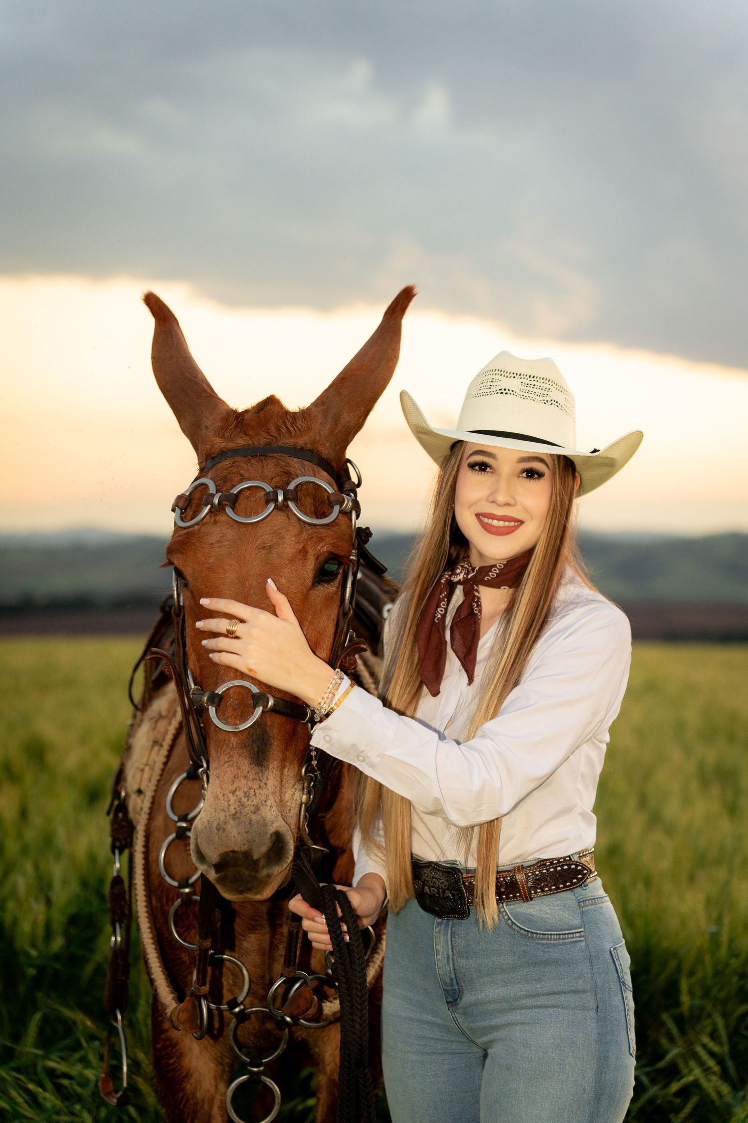 Young woman in cowboy hat and jeans smiles beside a brown horse in a green field at sunset.