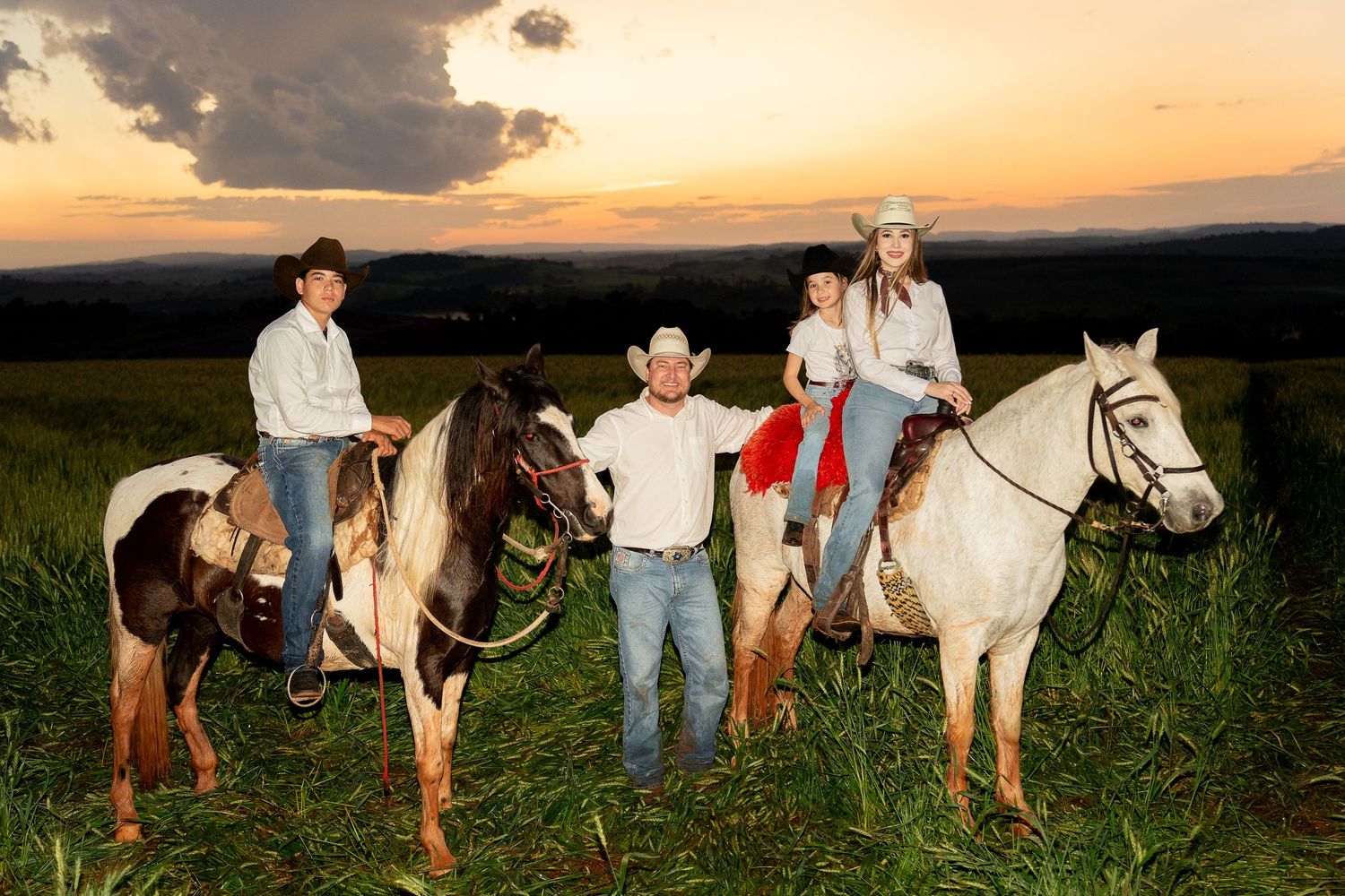 Family in cowboy hats posing with horses in a green field at sunset, wearing western attire.