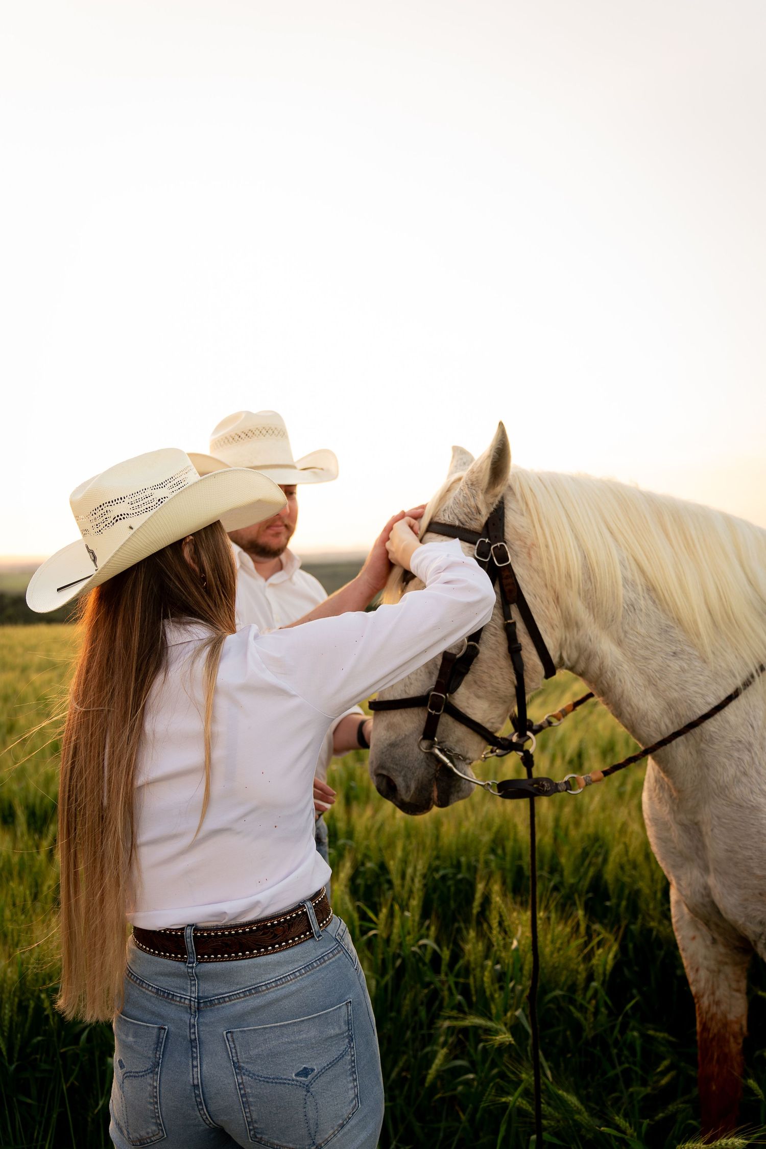 Woman in cowboy hat and white shirt petting a white horse in a sunlit green field.
