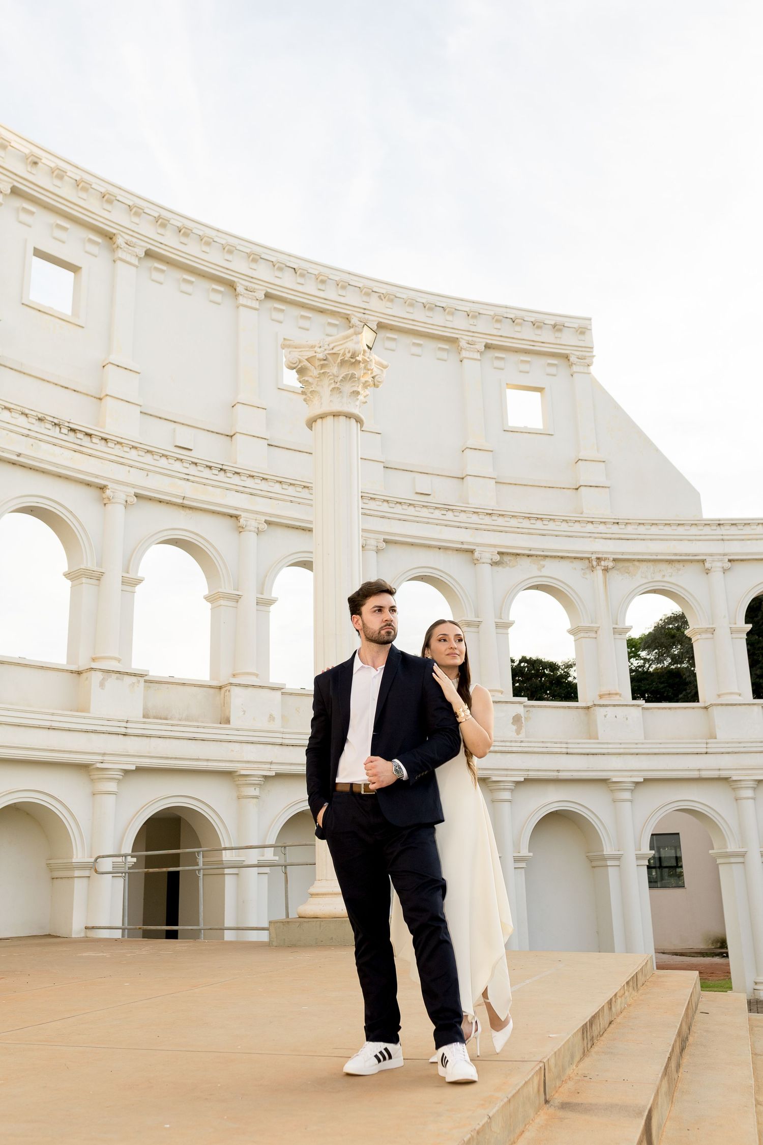 Elegant couple posing in front of a white Roman Colosseum replica, man in dark suit, woman in white dress.