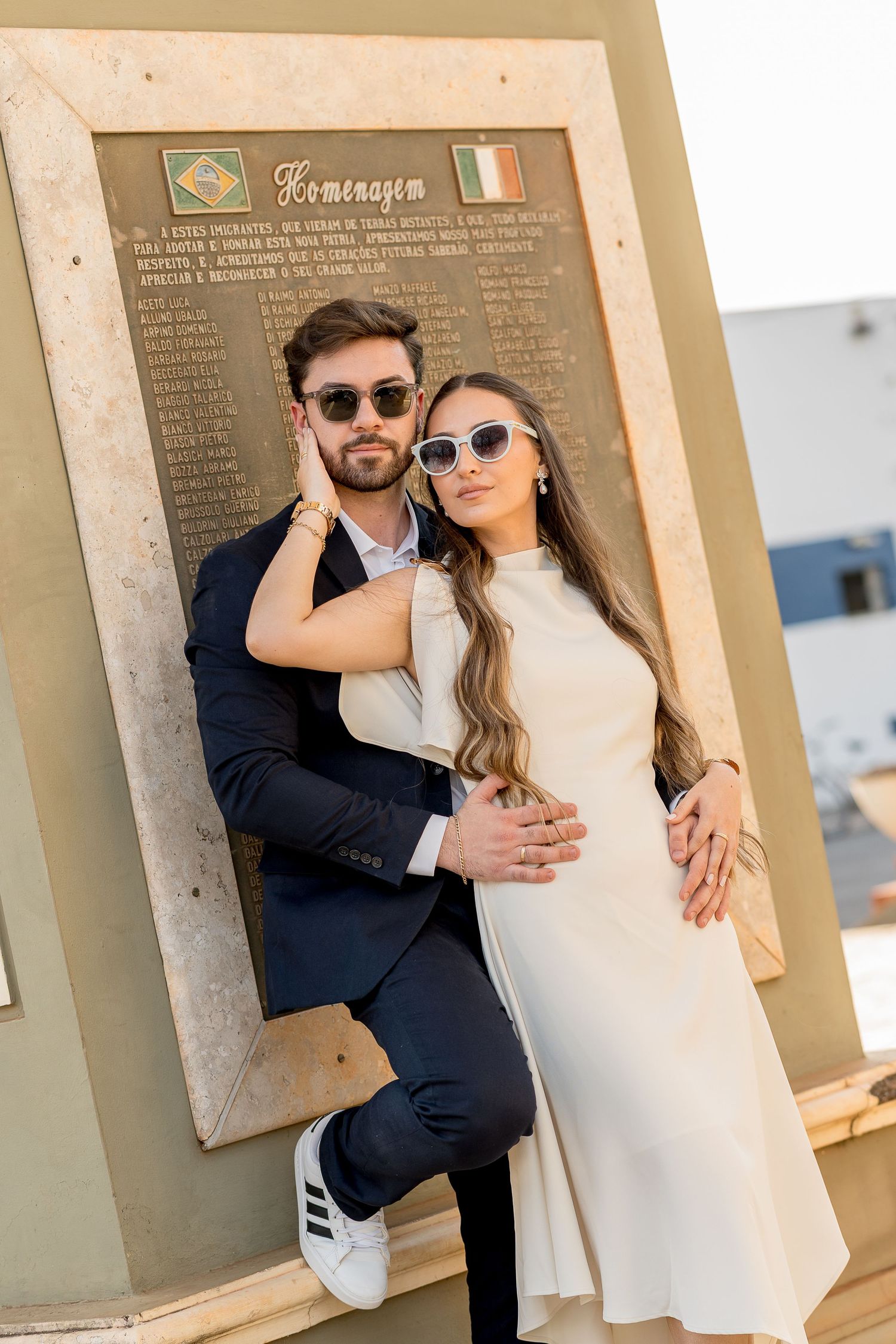 Stylish couple in sunglasses posing by a memorial plaque with Brazilian and Irish flags outdoors.