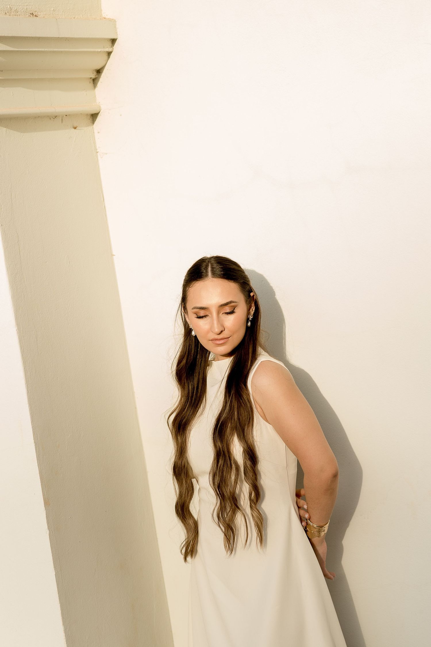 Woman in white sleeveless dress with long wavy hair leans against white wall near column, eyes downcast.