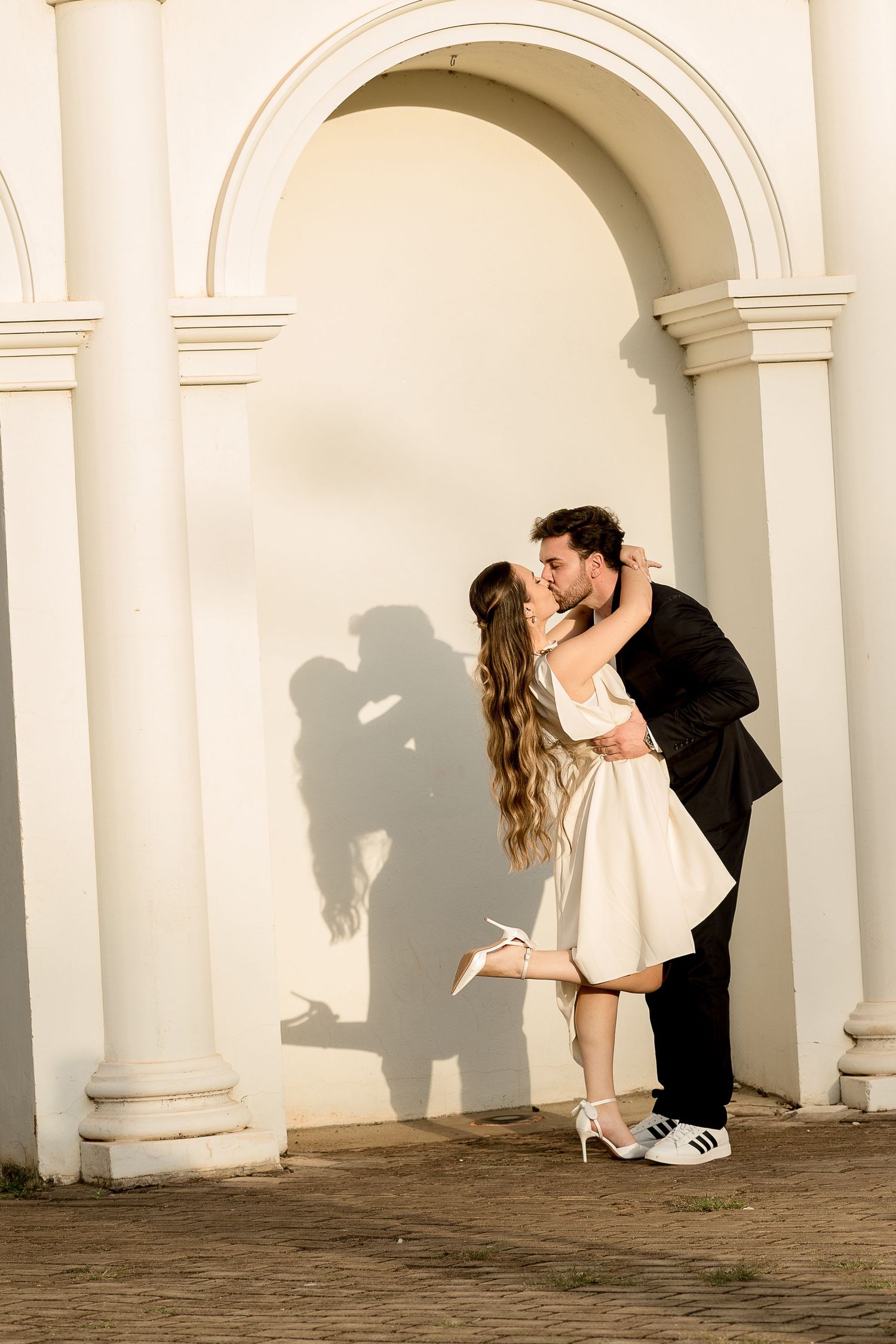 Romantic couple kissing under a white arch, casting a shadow on the wall, bride in white dress and groom in black suit.