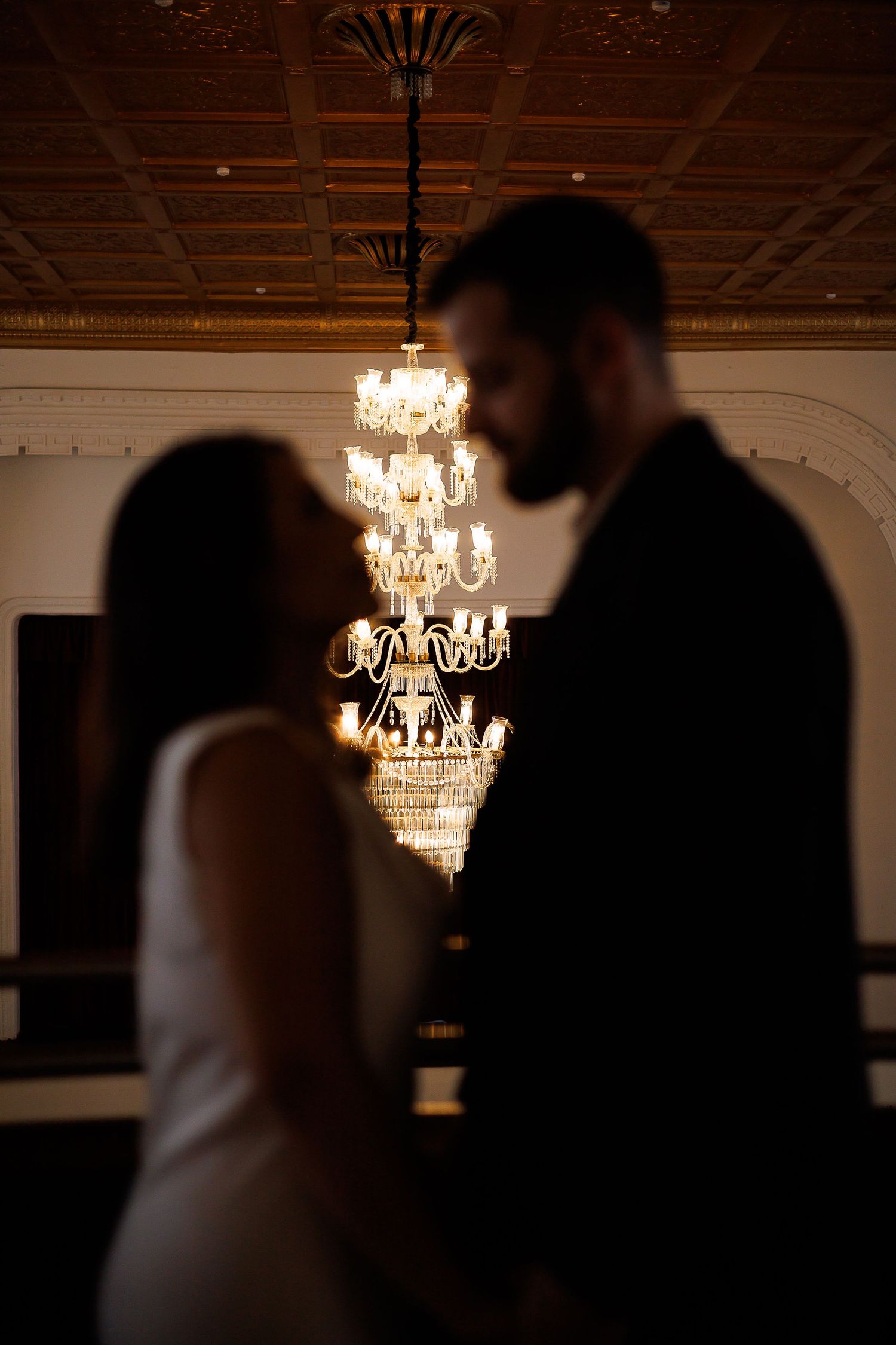 Couple sharing romantic moment in elegant ballroom with glowing crystal chandelier backdrop.