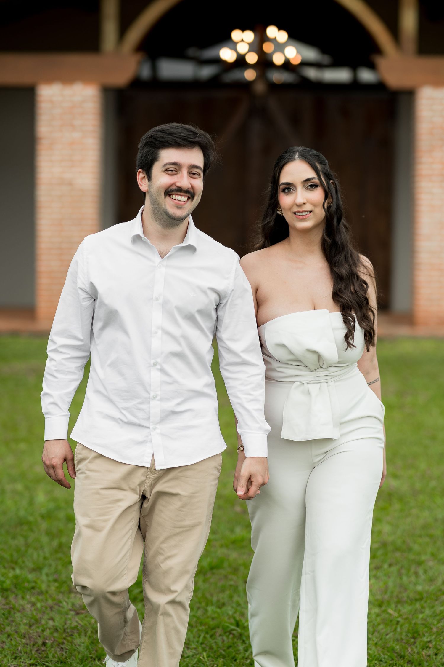 A smiling couple in white outfits holds hands while walking outdoors near a rustic barn venue.