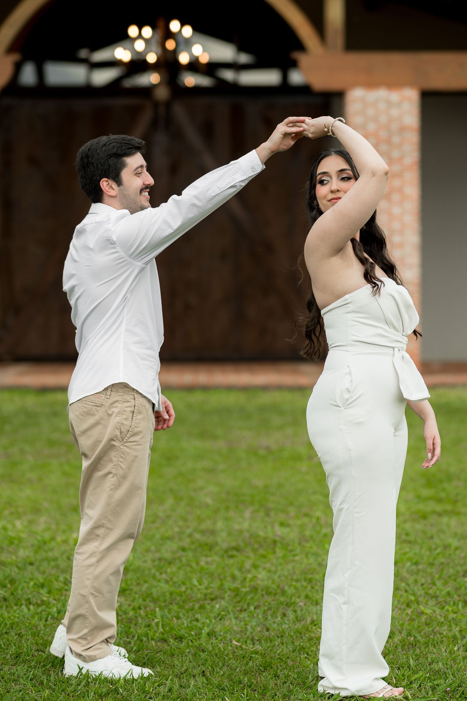 Couple dancing outdoors on green grass, man in white shirt and khaki pants spinning woman in elegant white outfit.