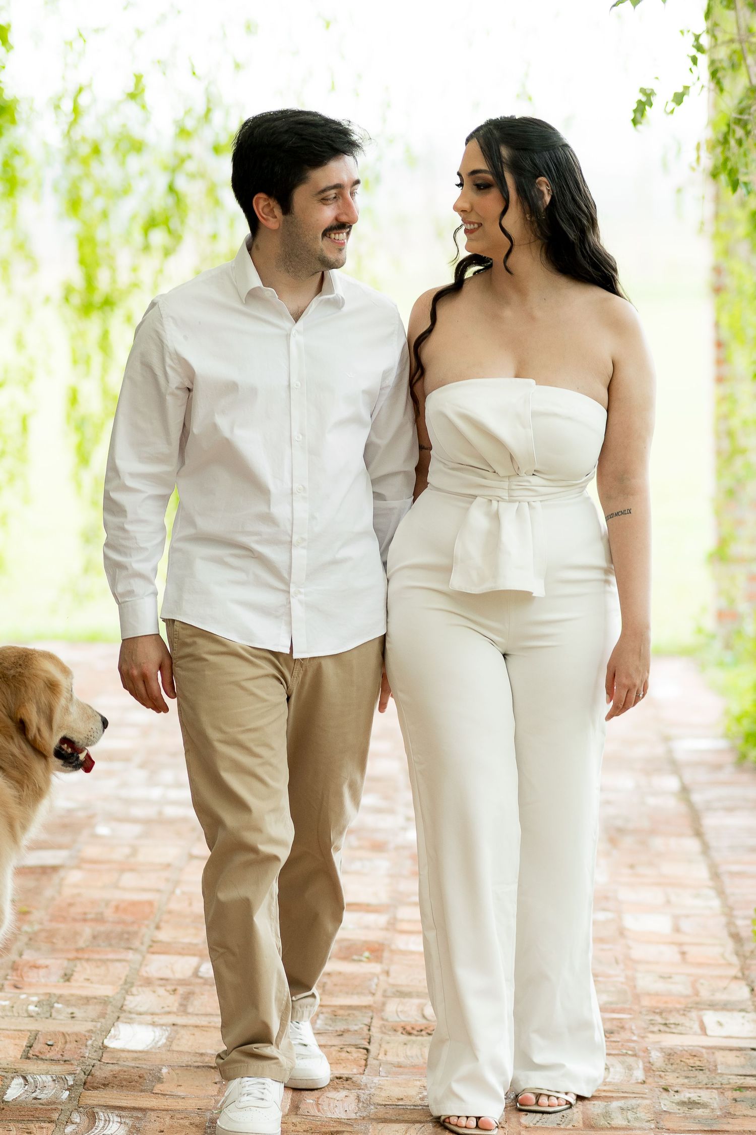 A smiling couple in white outfits walks together on a brick path surrounded by lush greenery, with a golden dog nearby.