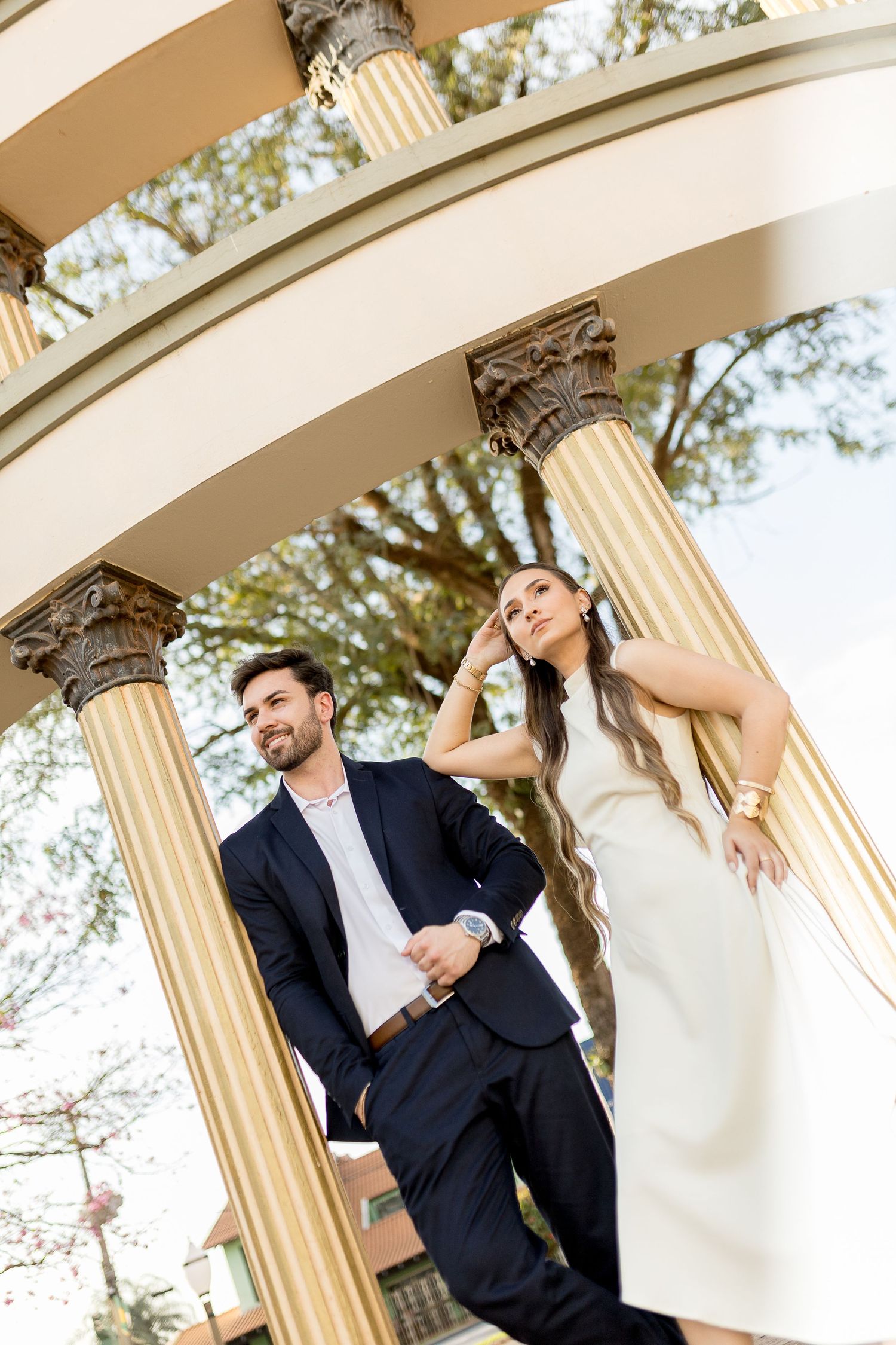 Elegant couple posing under a classical gazebo with gold columns, man in navy suit and woman in white dress.