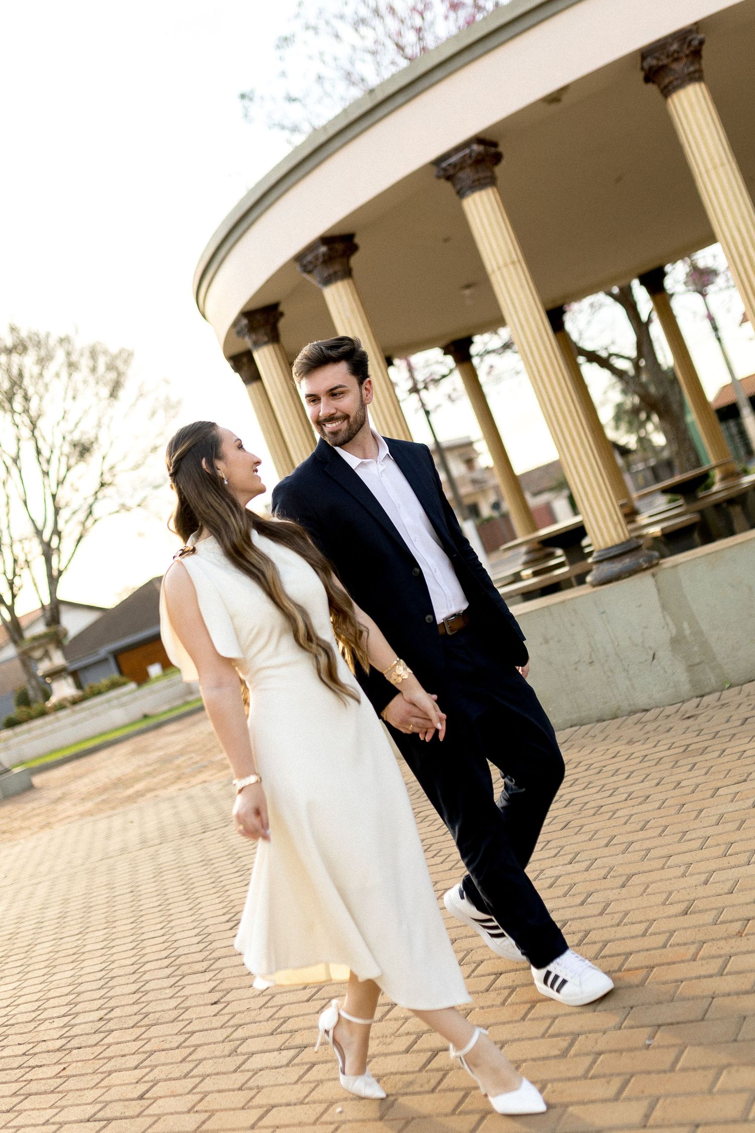 Couple holding hands in front of a classical rotunda, woman in white dress and man in navy suit smiling at each other.