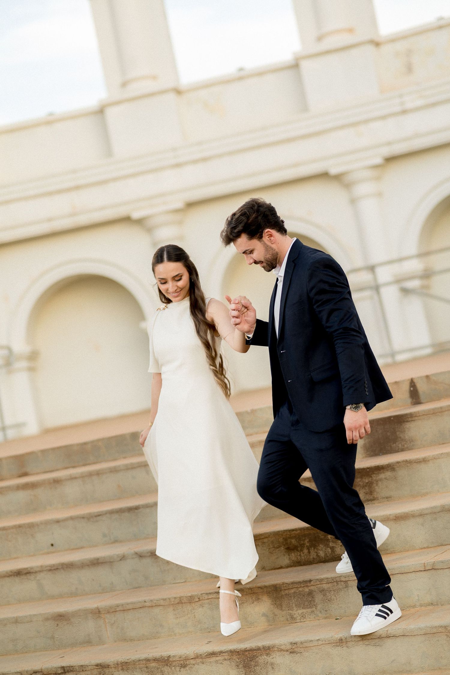 Elegant couple in wedding attire holding hands while descending grand stone steps of a classical building.