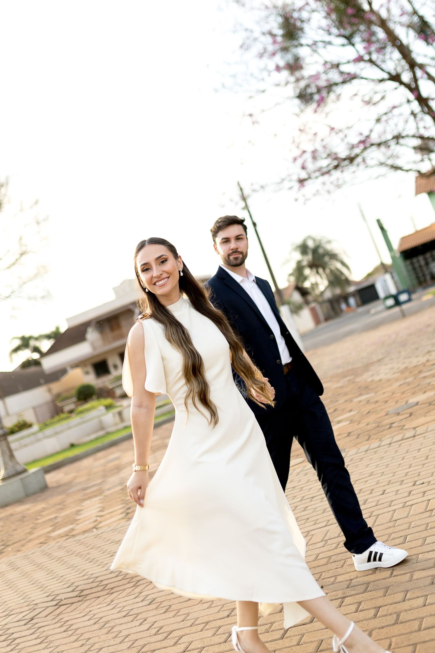 Smiling bride in white dress walks with groom in navy suit on brick plaza with blooming trees in background.