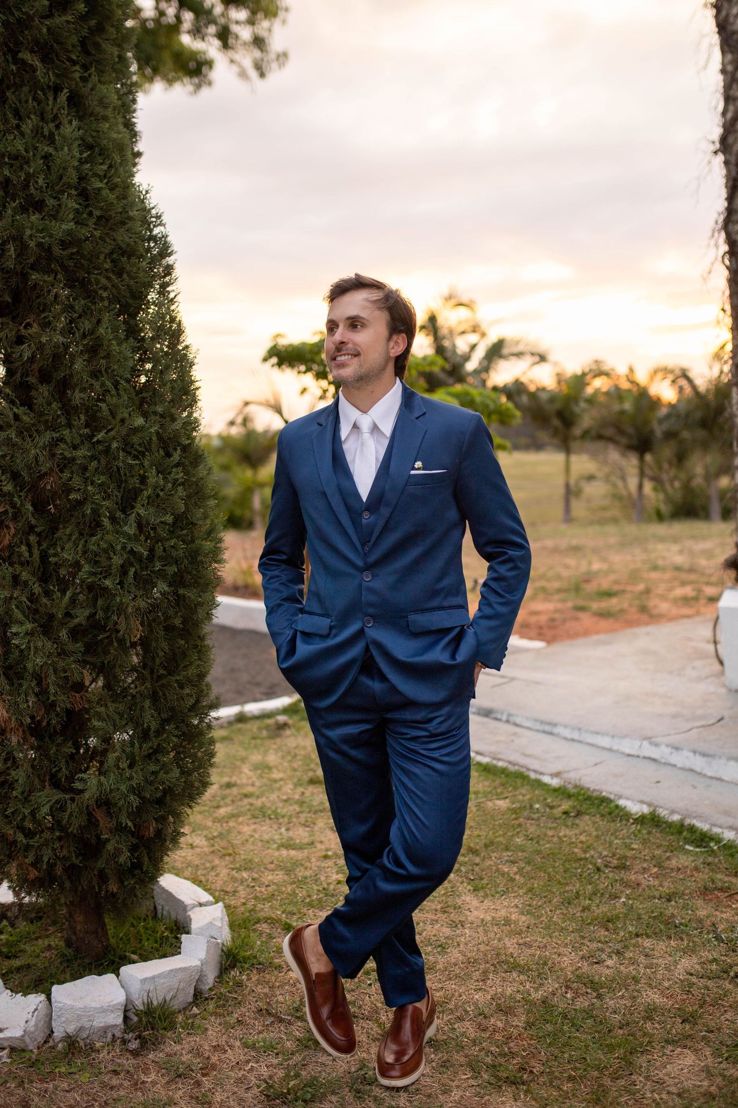 Man in navy blue three-piece suit with white tie and brown shoes standing outdoors at sunset near cypress trees.