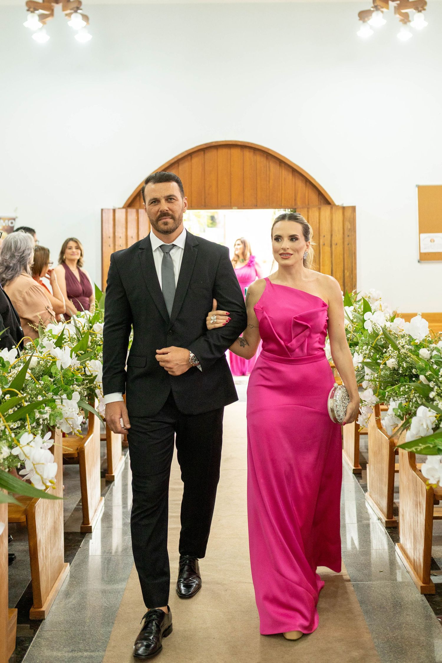 A couple walks down a church aisle, man in black suit and woman in pink gown, flanked by floral arrangements.