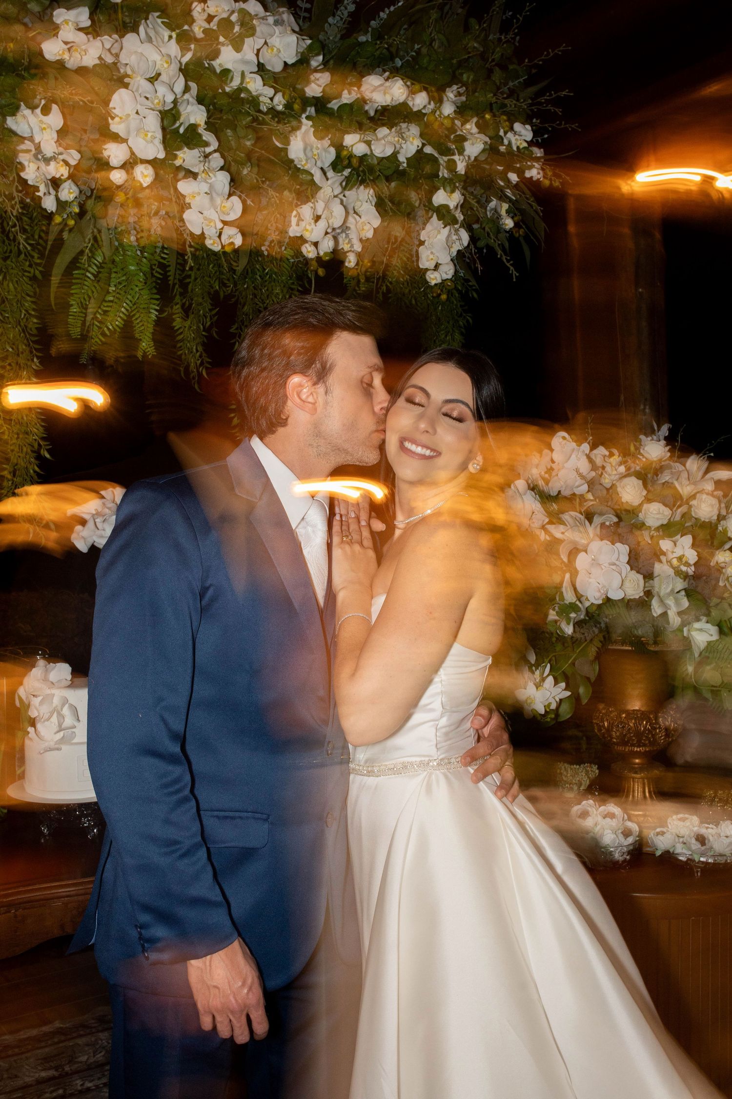 A groom in a blue suit kisses his smiling bride in a white gown at their elegant floral wedding reception.