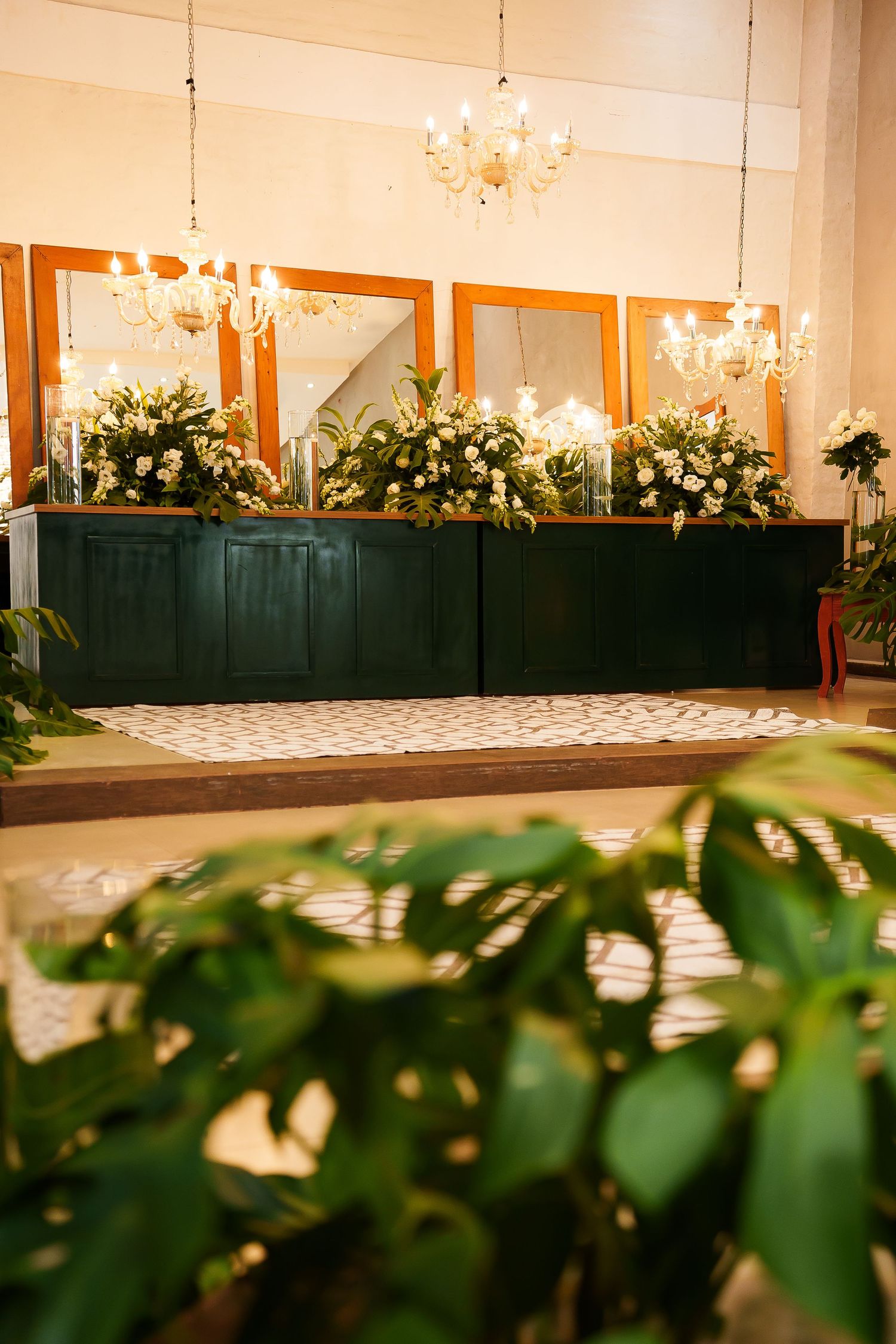 Elegant event hall with chandelier lighting, green buffet table, floral arrangements, and mirrors reflecting white flowers.