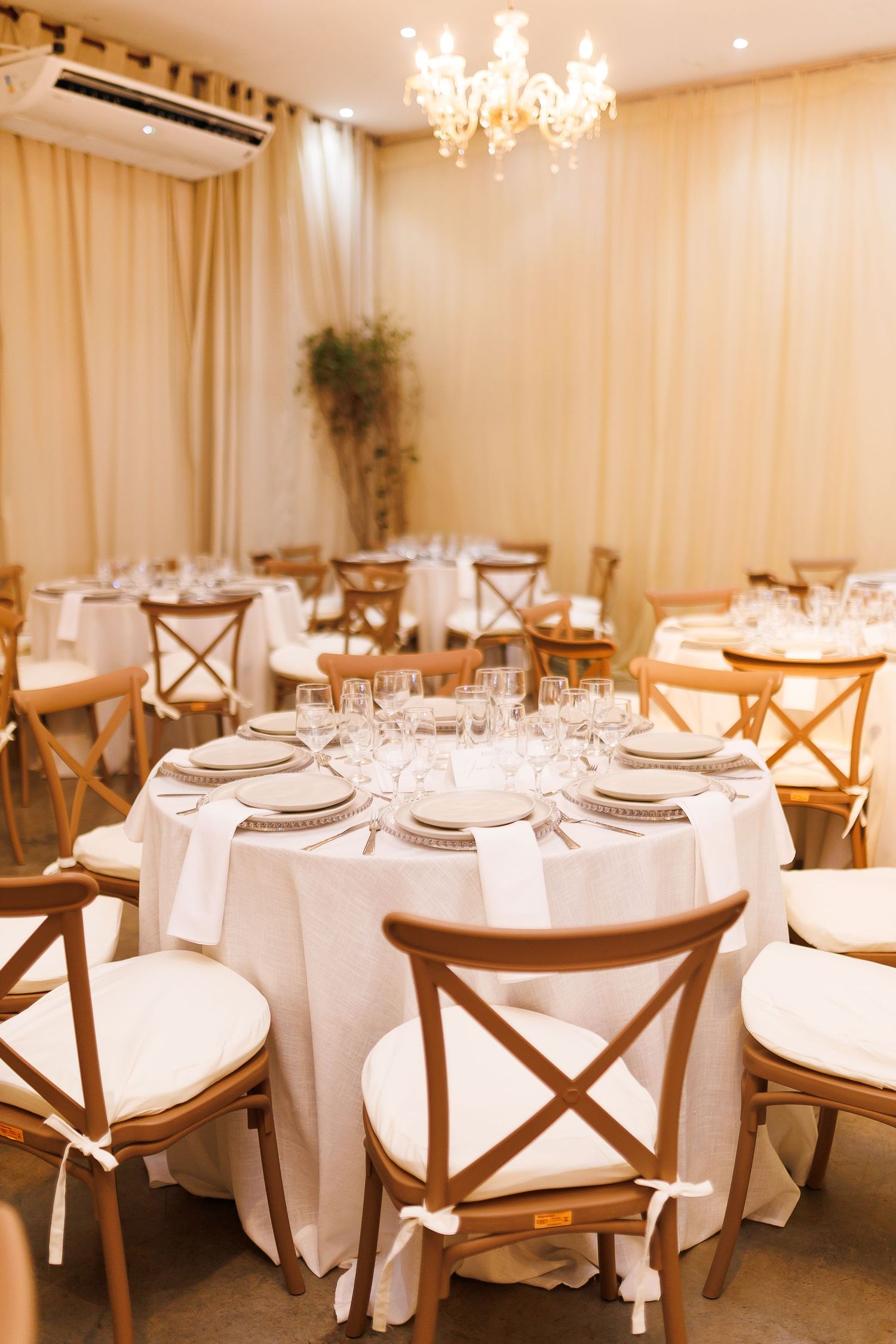 Elegant banquet hall with round tables, white linens, wooden cross-back chairs, crystal glasses, and a chandelier.