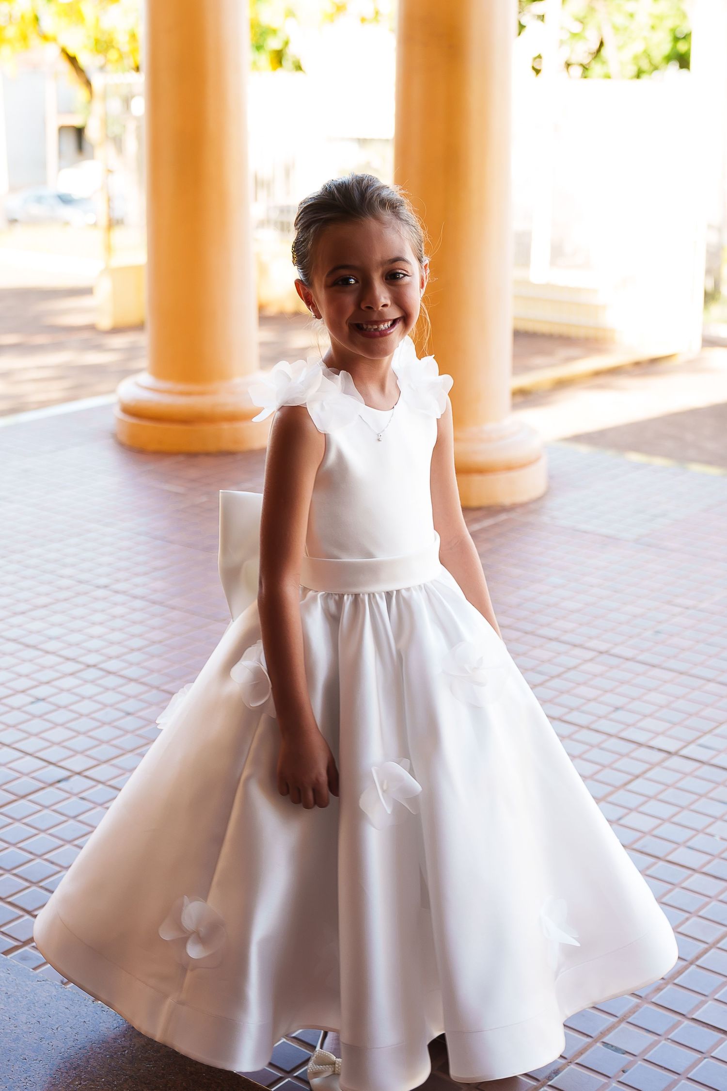 Smiling young girl in white flower girl dress with floral details posing between yellow columns.
