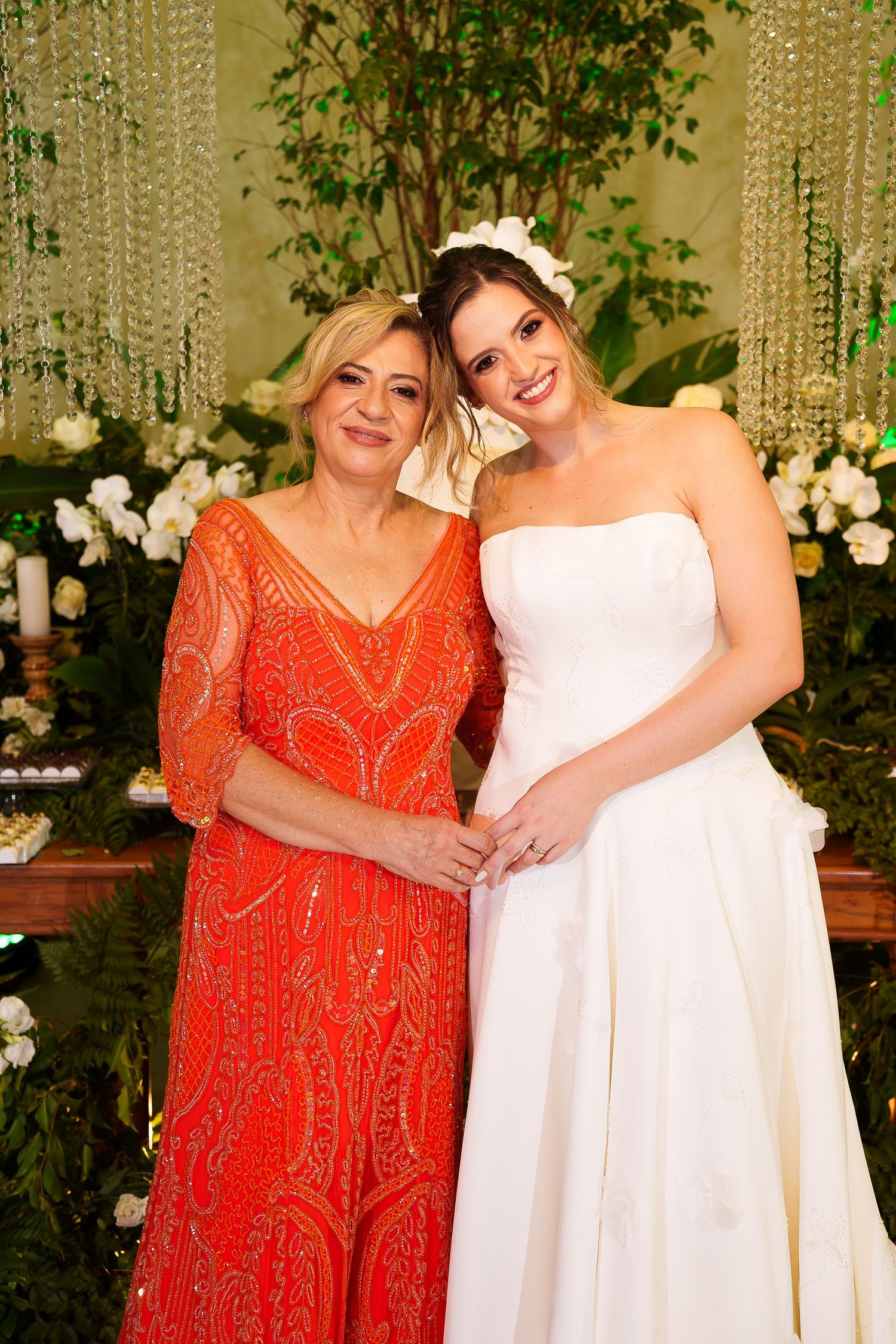 A bride in white strapless gown poses with an older woman in an orange beaded dress at a wedding reception.