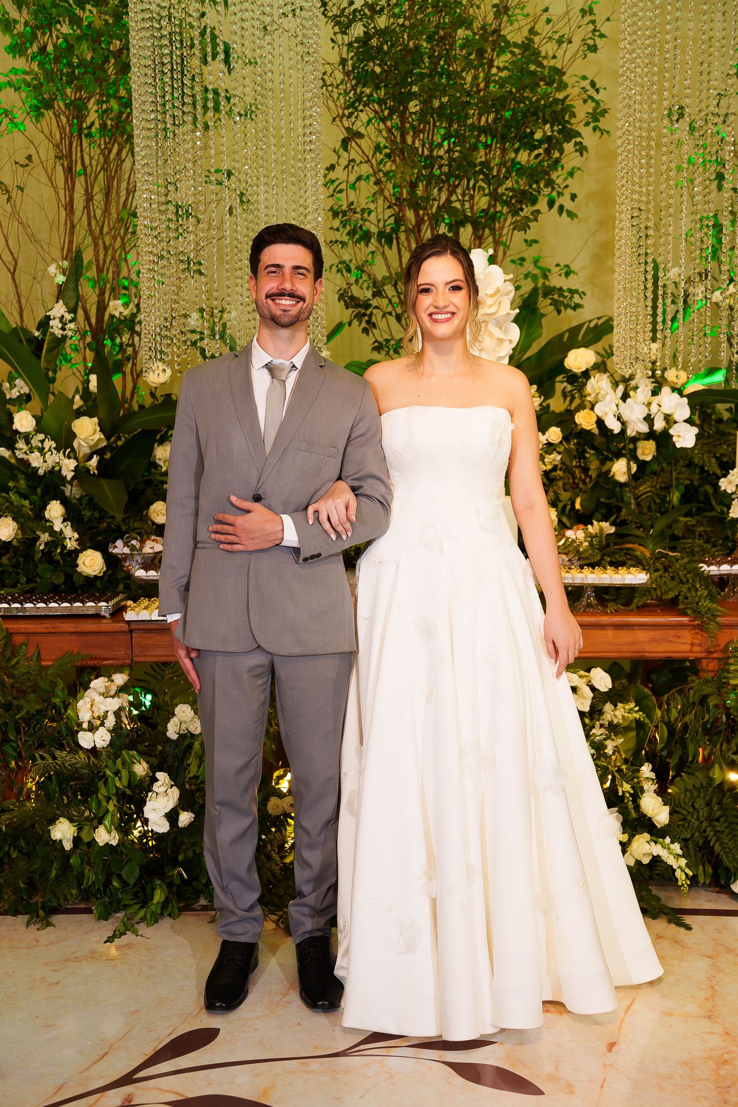 A smiling bride in a white strapless gown and groom in a gray suit pose together before a lush floral backdrop.