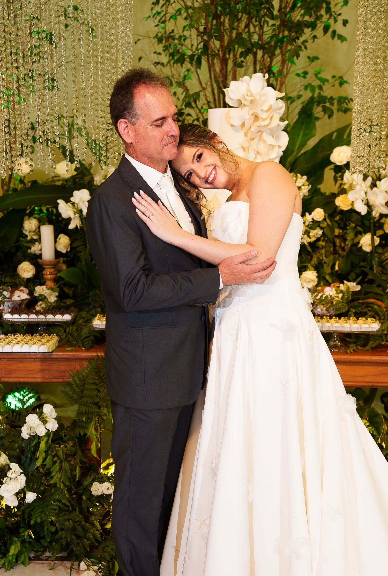 Bride in strapless ball gown embracing man in dark suit near floral backdrop at wedding reception.