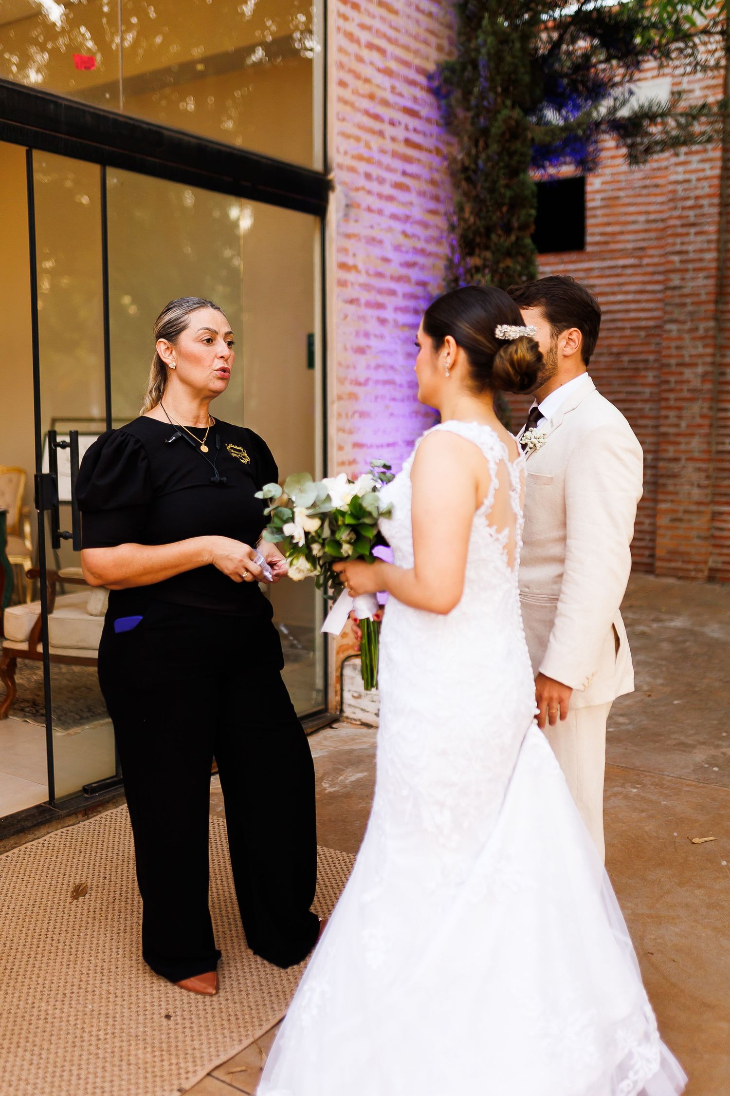 Wedding officiant in black speaks with bride in white lace gown and groom in beige suit outdoors.