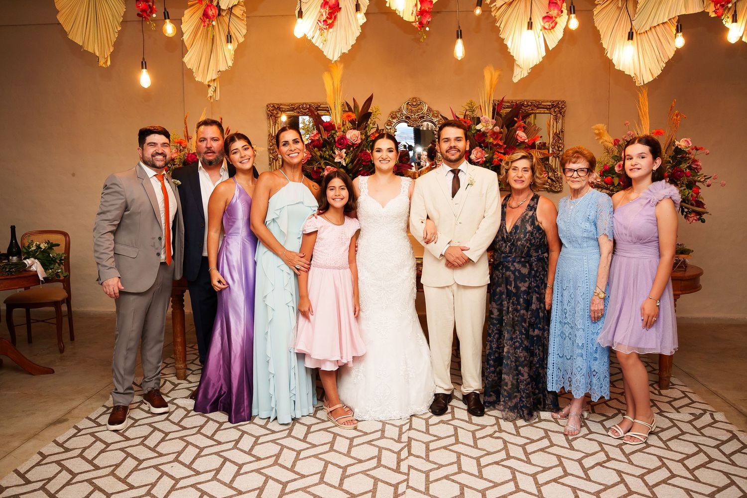 Wedding party posing together in elegant venue with floral decor, bride in white gown surrounded by family in colorful dresses.