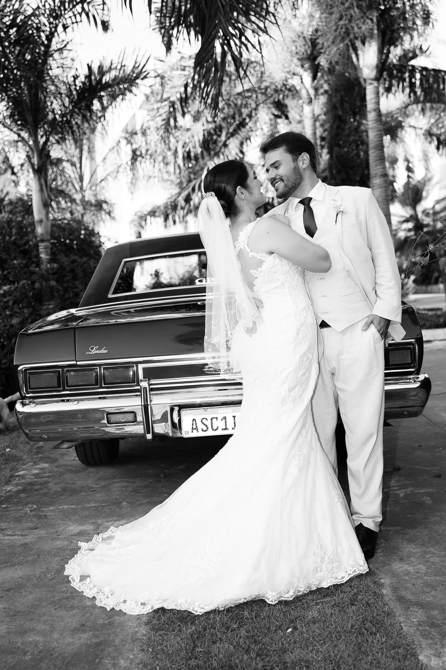 Black and white wedding photo of bride and groom embracing by a classic vintage car surrounded by palm trees.