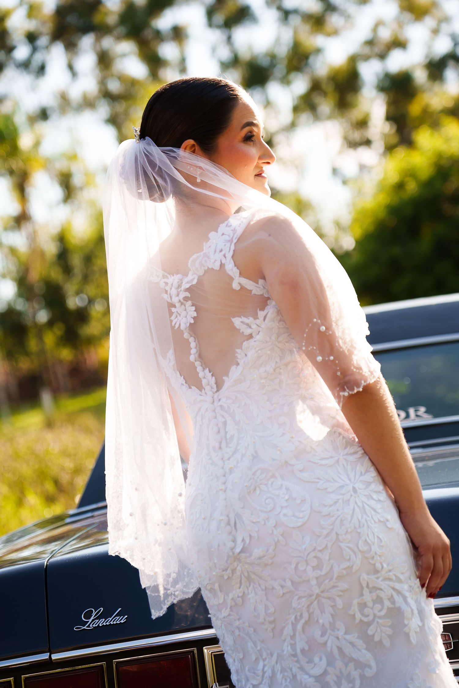 Bride in lace wedding dress and veil poses beside a vintage black Landau car outdoors.