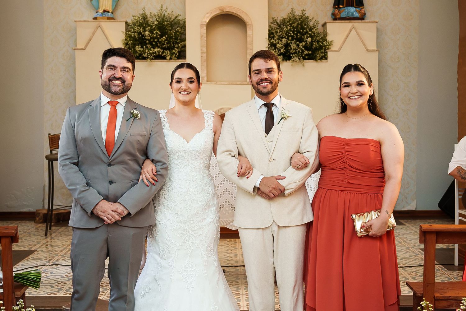 Wedding party posing in church aisle, bride in white gown, groom in cream suit, groomsman in gray, bridesmaid in red dress.