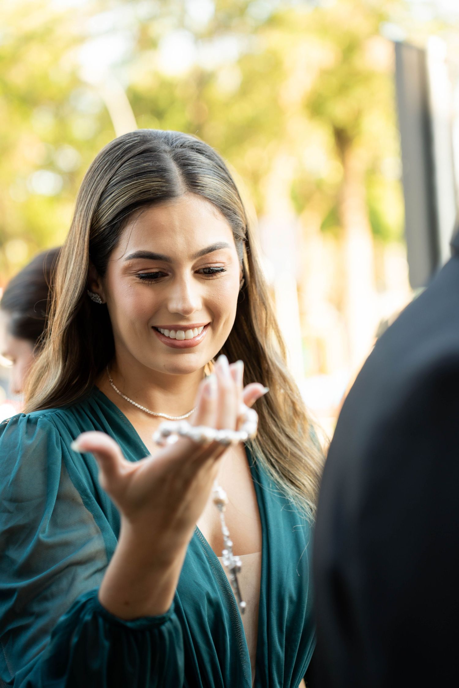 Smiling woman in teal dress examining jewelry outdoors at elegant event.