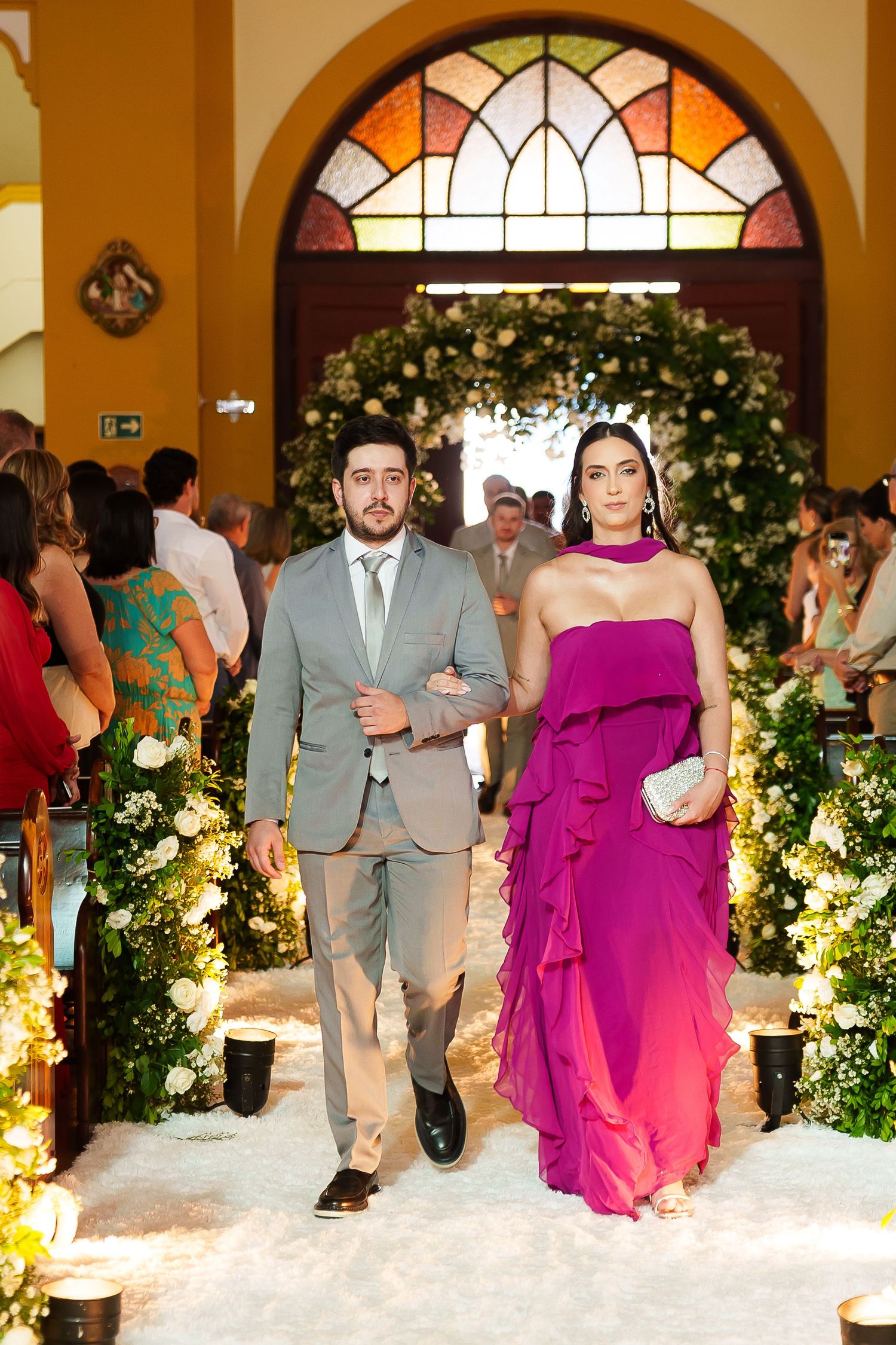 Couple walks down a floral-lined wedding aisle, man in gray suit, woman in vibrant magenta ruffled gown.