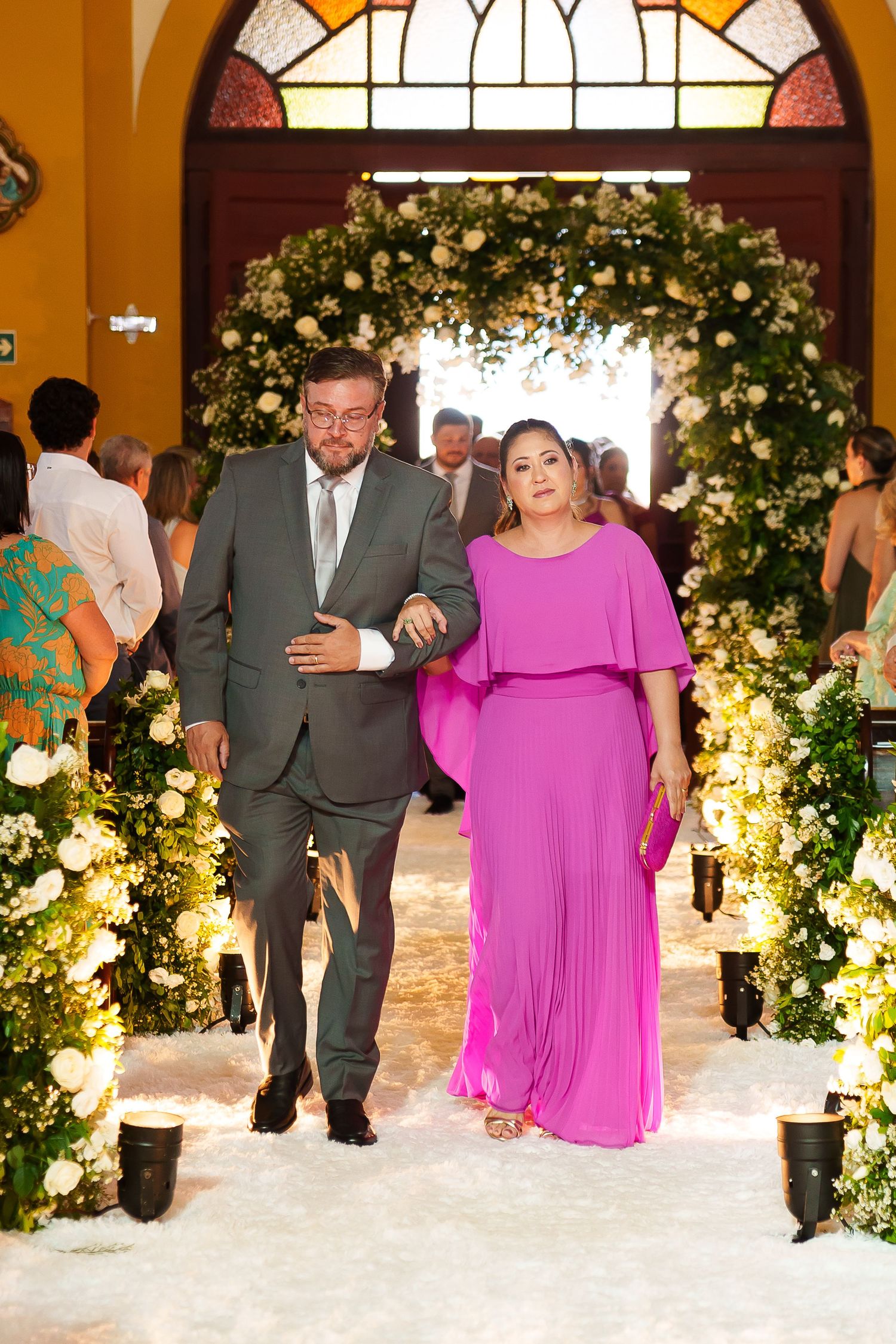 Couple walking down wedding aisle, man in gray suit, woman in pink gown, floral arch backdrop.