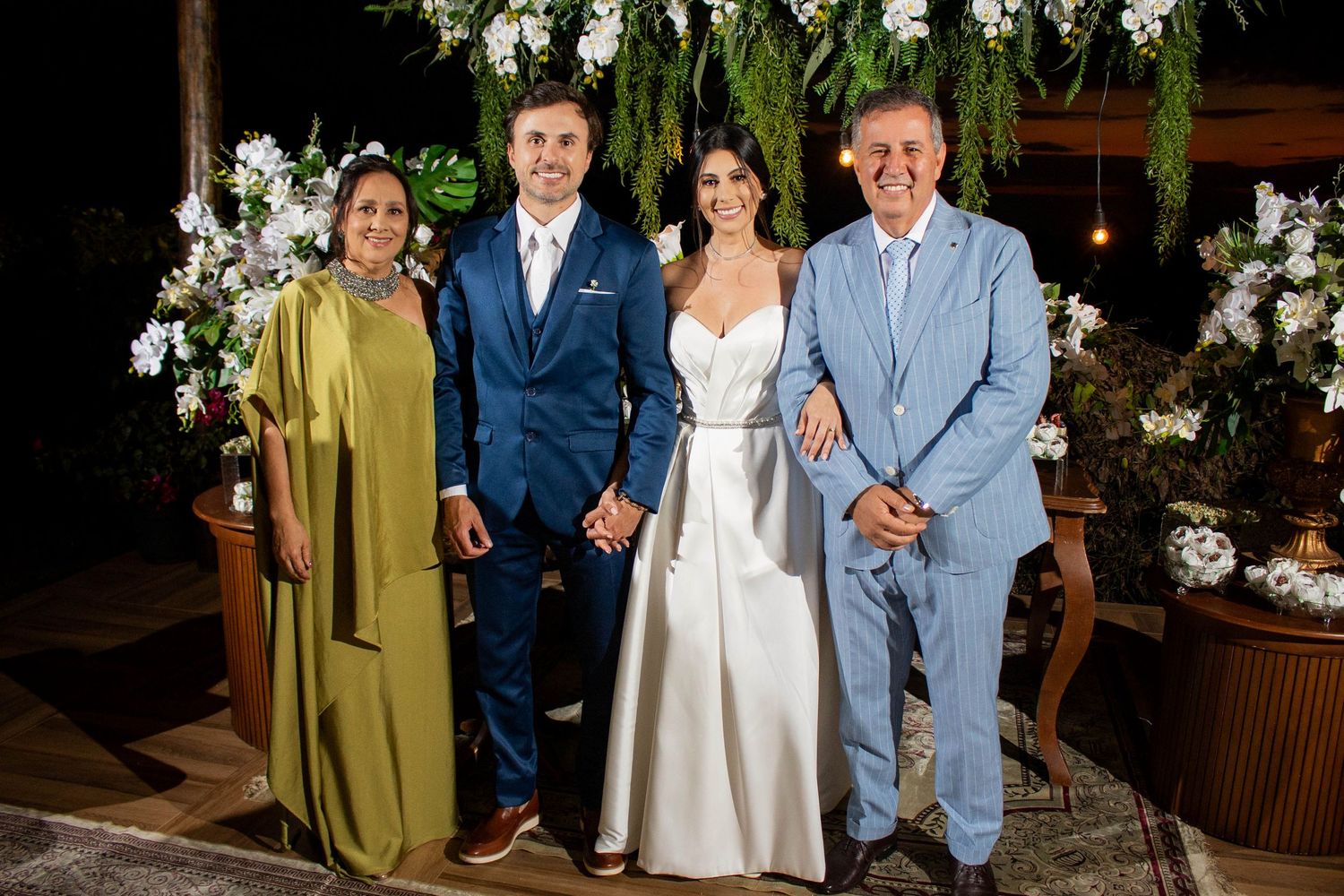 Bride and groom pose with family members in formal attire at an elegant outdoor wedding ceremony with floral decorations.