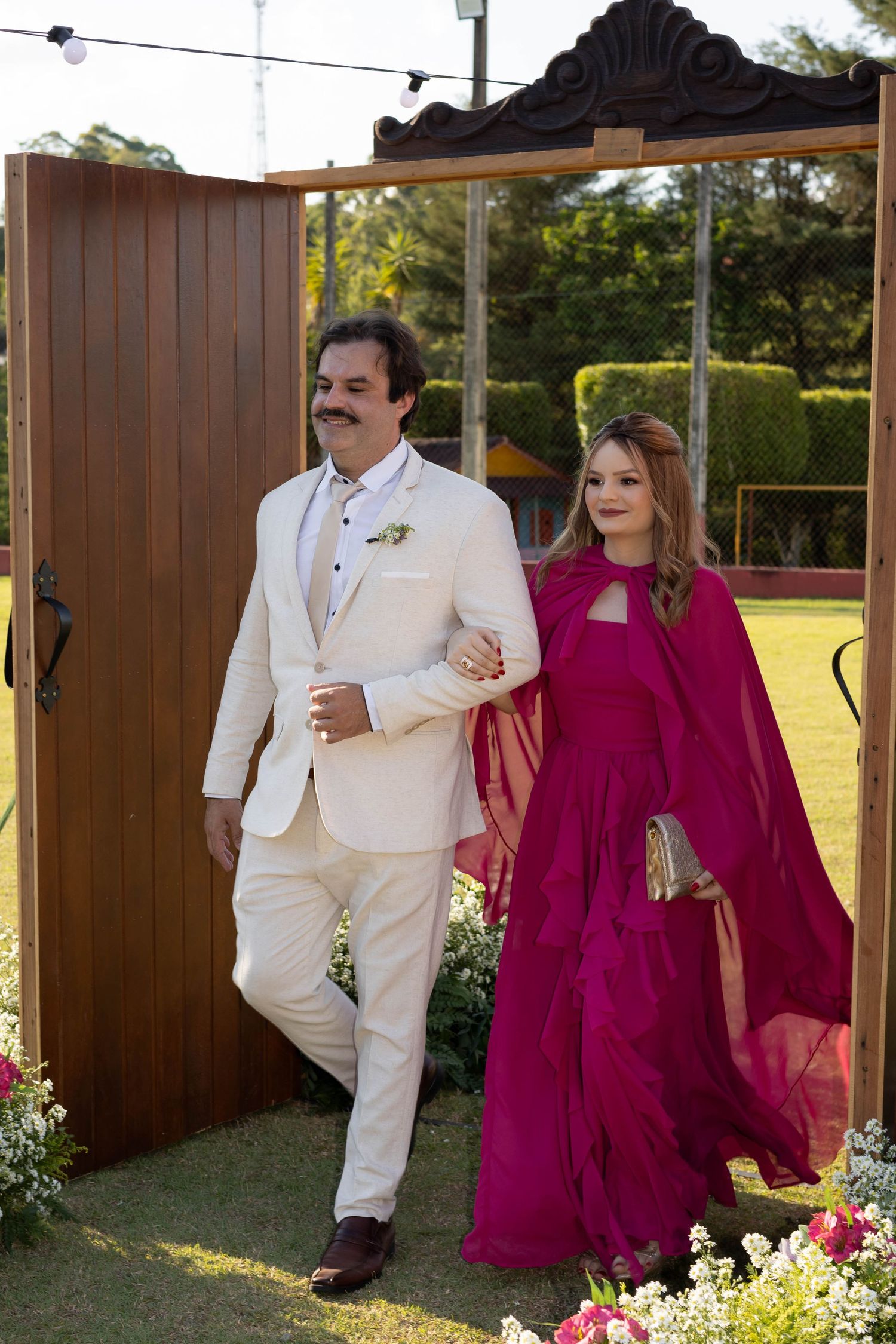 Elegant couple at outdoor wedding, man in cream suit, woman in magenta gown with cape, walking through wooden arch.