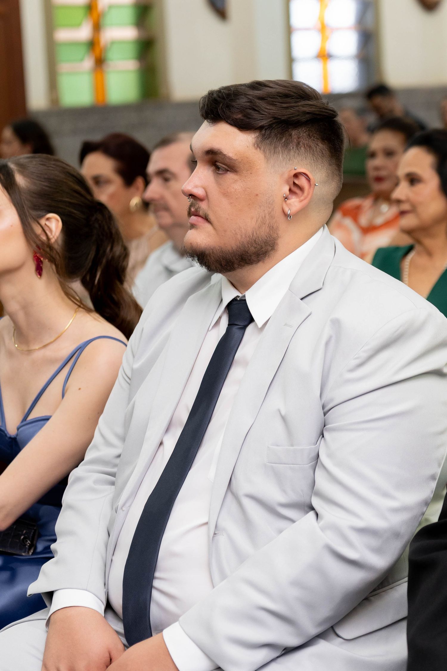Young man in white suit and black tie seated at a formal church ceremony event.
