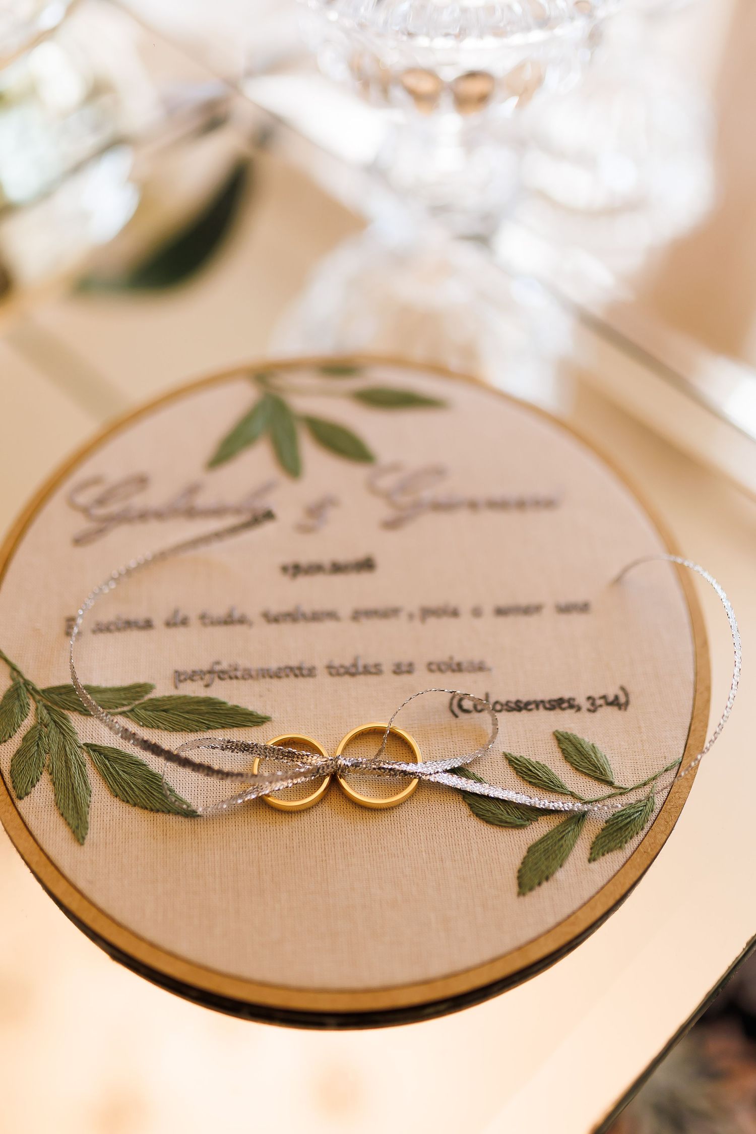 Two gold wedding rings rest on an embroidered hoop with green leaves and a romantic script quote for a wedding ceremony.