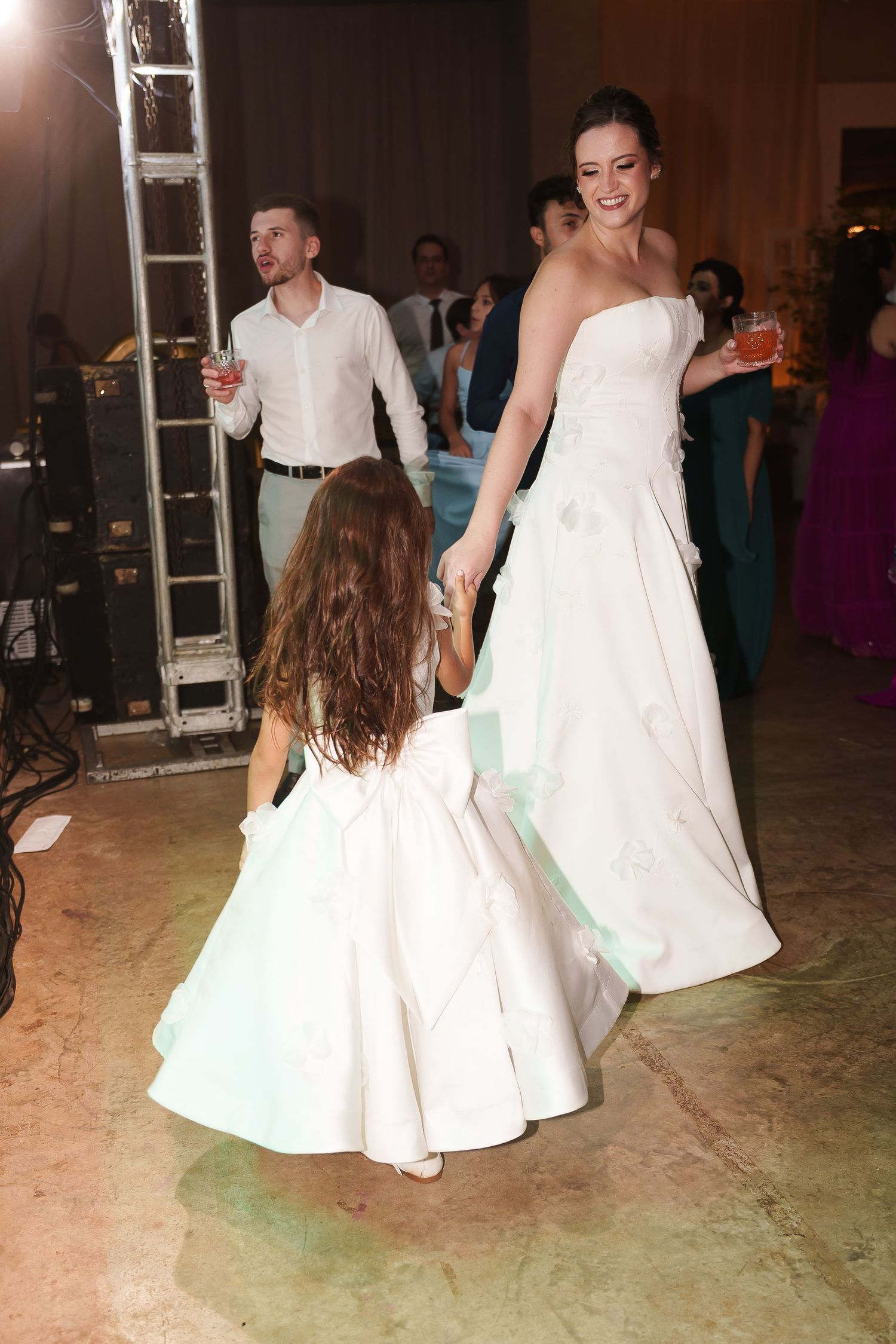 Bride in white gown dancing with flower girl at wedding reception while guests celebrate in background.