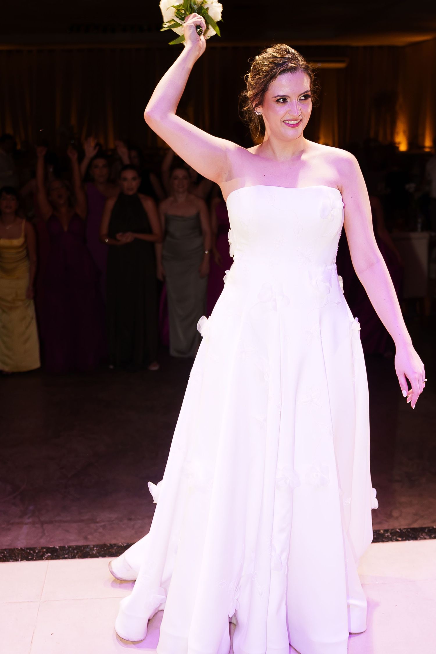 Bride in white strapless gown raises bouquet during wedding reception bouquet toss with guests in background.