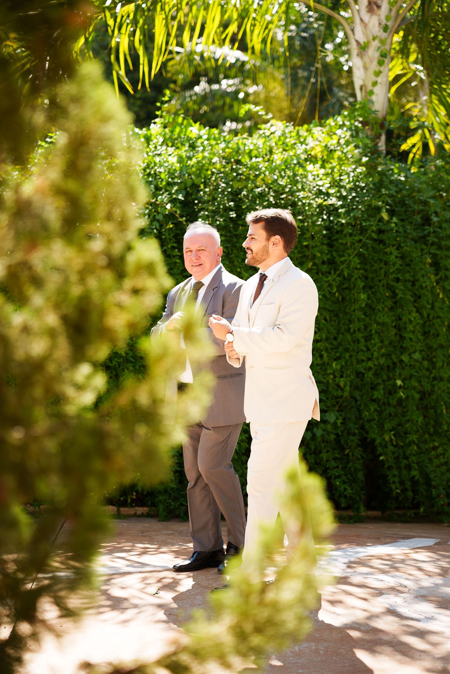 Two men in suits conversing outdoors in a lush green garden setting on a sunny day.