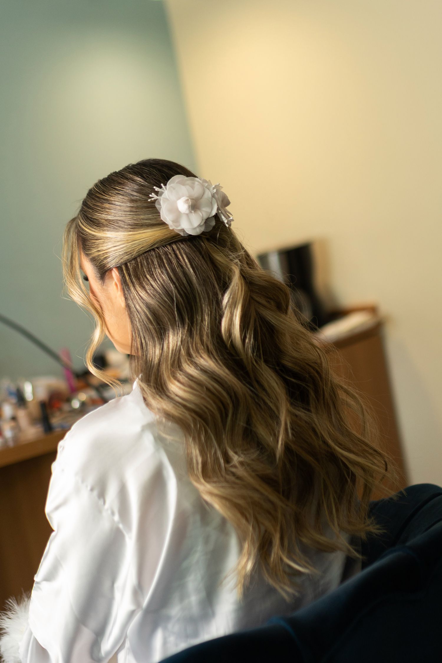 Bride with half-up wavy hairstyle adorned with a white floral hair accessory, wearing a white robe.