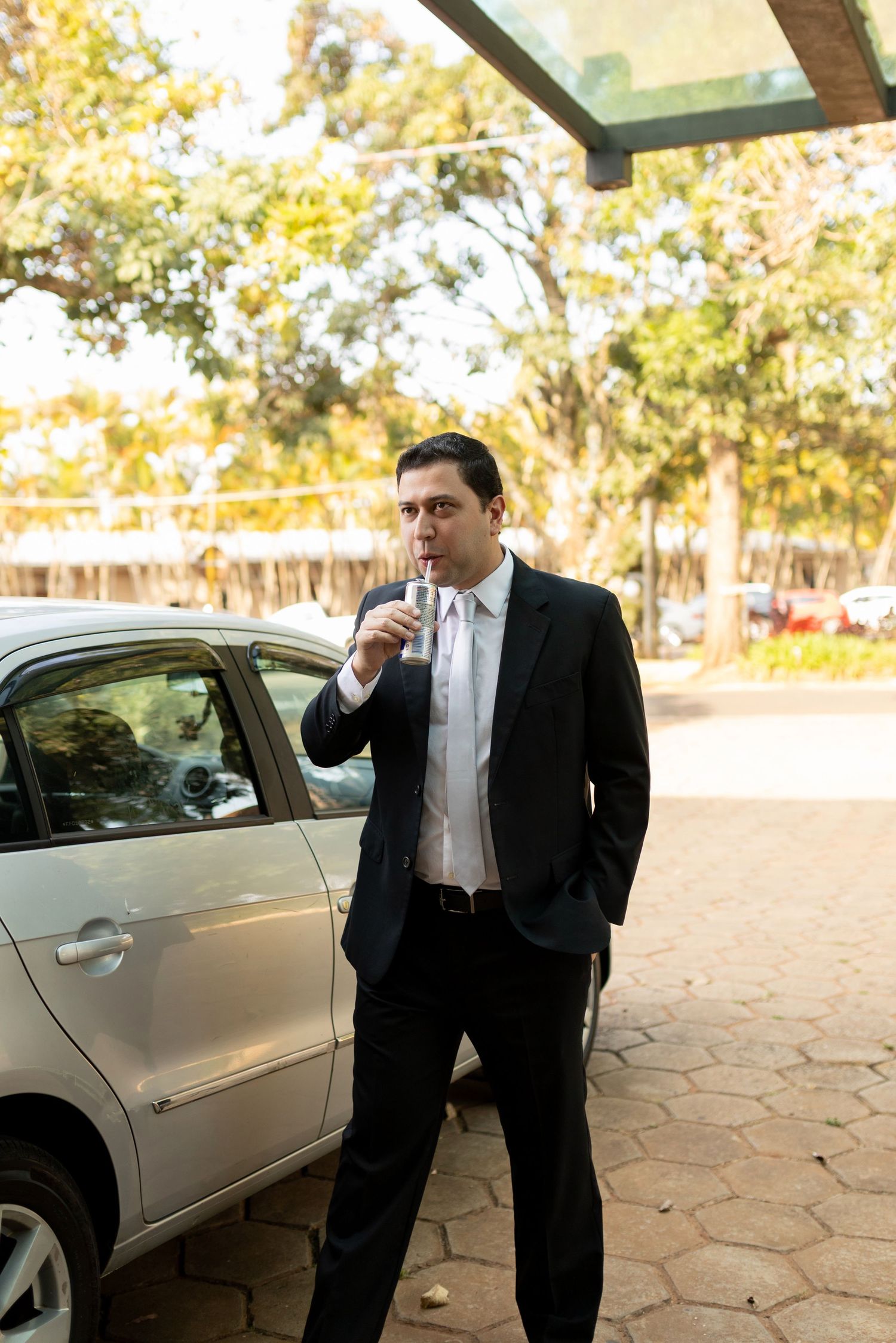 Businessman in black suit smoking a cigarette beside a silver car in an outdoor parking area with trees.