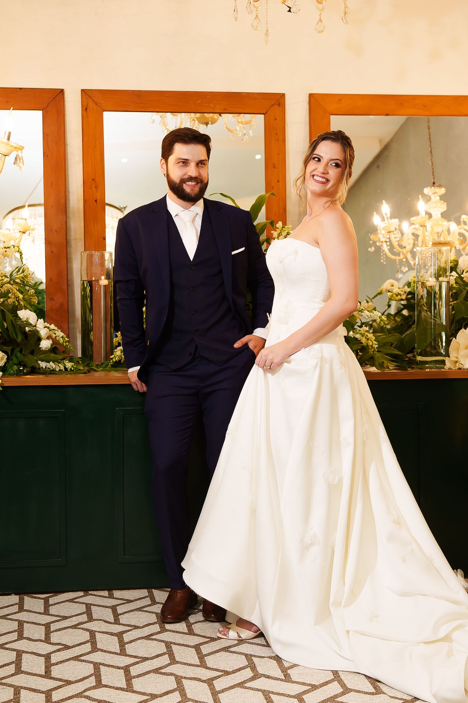 A smiling bride in a white gown and groom in a navy suit pose together in an elegant chandelier-lit venue.