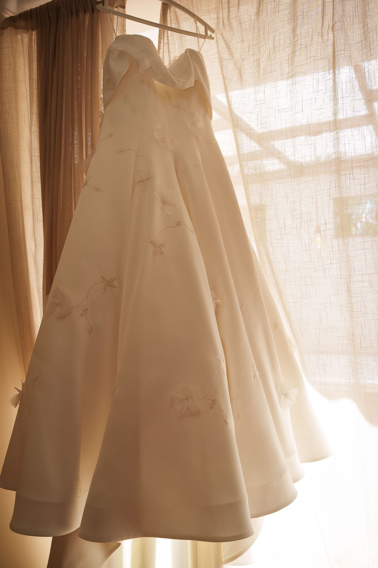 Elegant strapless ivory wedding gown with floral embroidery hanging near a sunlit sheer curtained window.