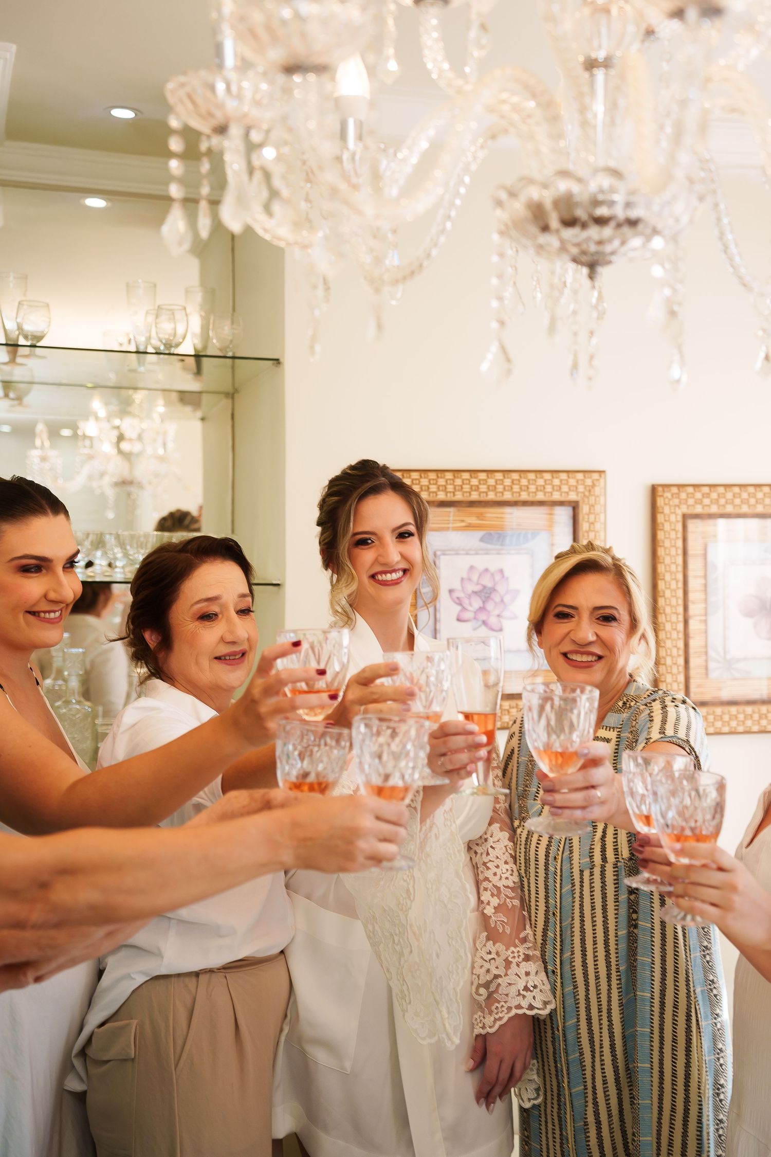 Bride and bridesmaids toasting with rose wine glasses under crystal chandeliers during wedding preparations.