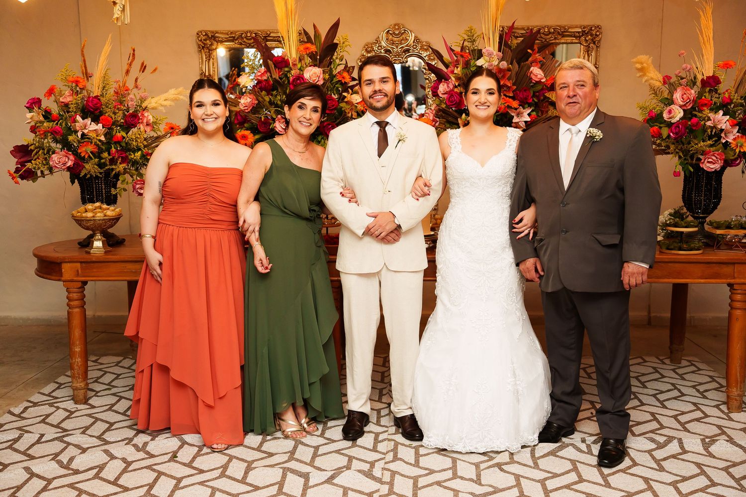 Wedding party poses indoors with bride in white gown, groom in ivory suit, and guests in orange and green dresses.